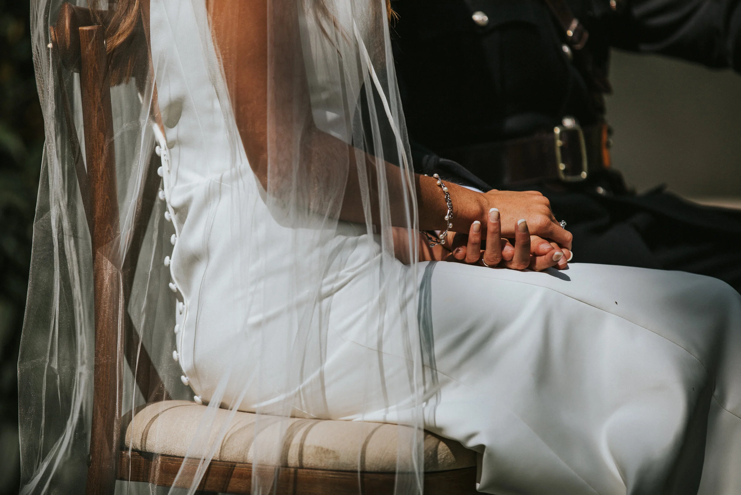 Close-up of two people holding hands, with one person wearing a white dress and the other in a black shirt, sitting on a wooden chair with a sheer curtain in the foreground.