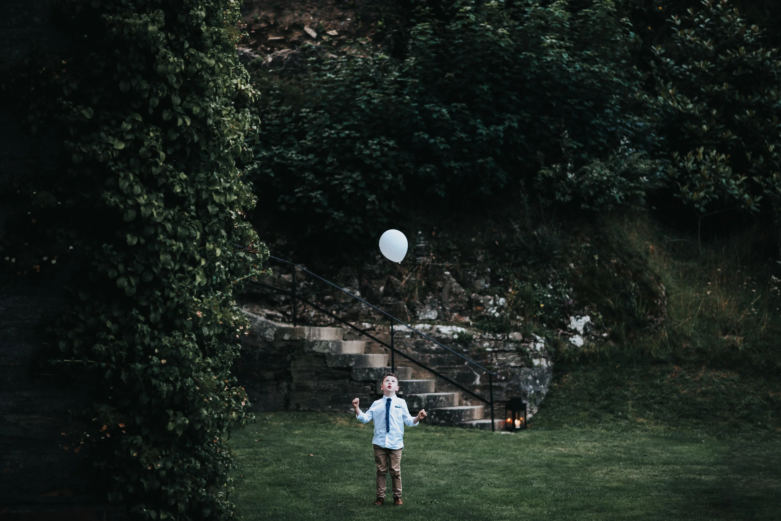 A young boy in a white shirt, blue tie, and brown pants is outdoors at dusk, playing with a white balloon in a backyard with green grass, large bushes, and stone steps with black railing leading up a hill.
