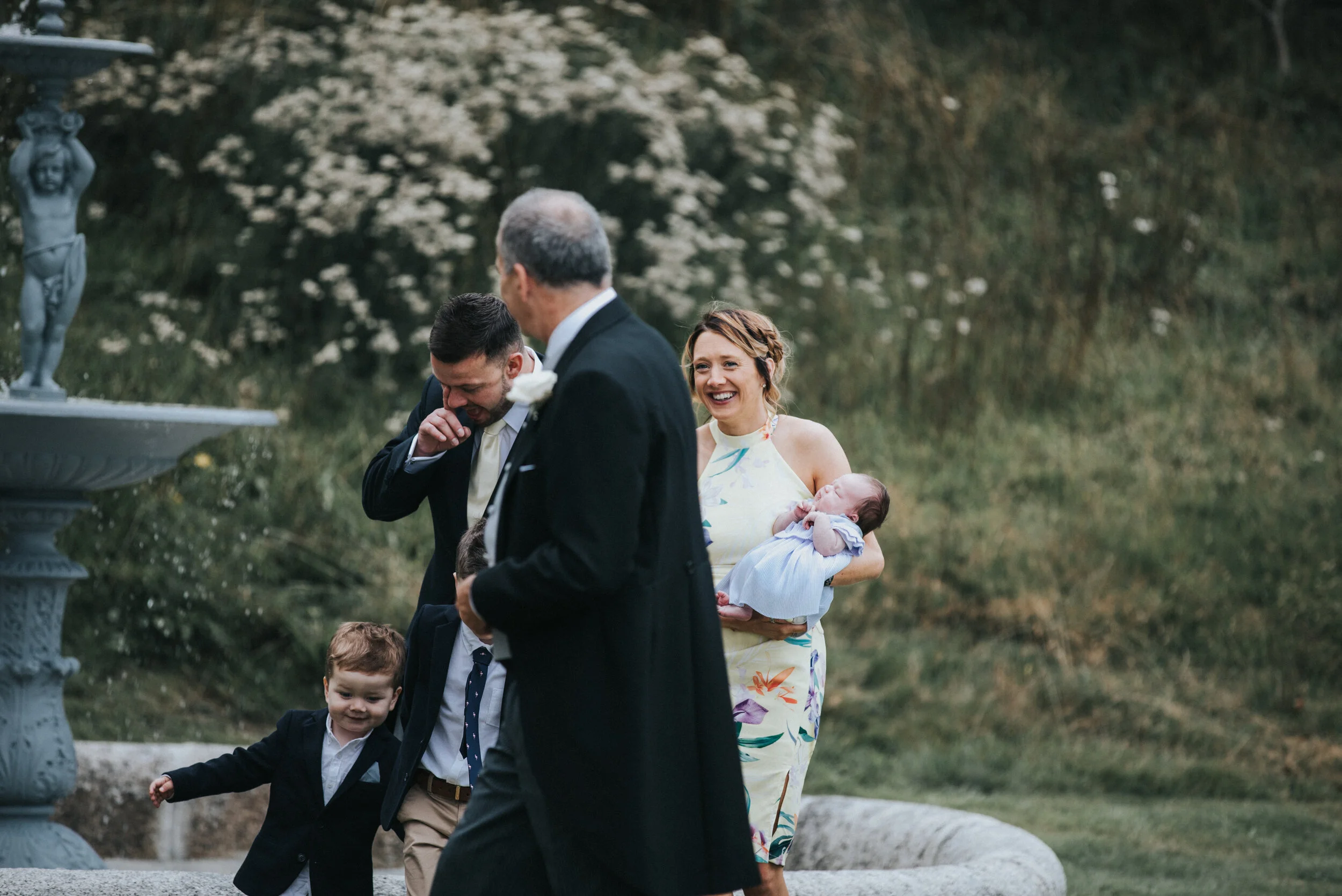 A group of people, including a woman holding a baby, two young boys, and an older man, are outdoors near a fountain and bushes. They are dressed in formal attire, suggesting a special occasion or celebration.