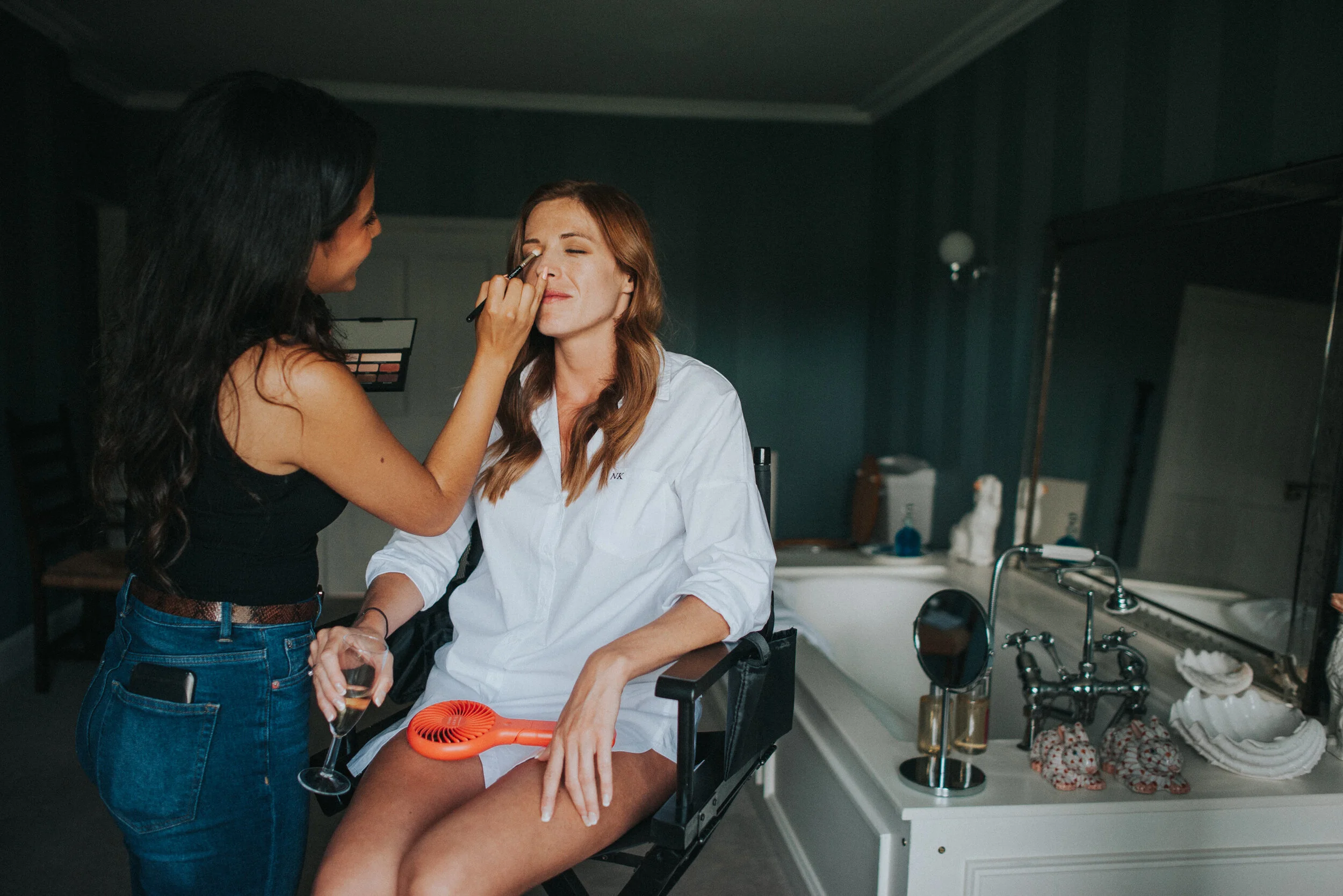 Makeup artist applying makeup to woman sitting in a makeup chair in a bathroom.