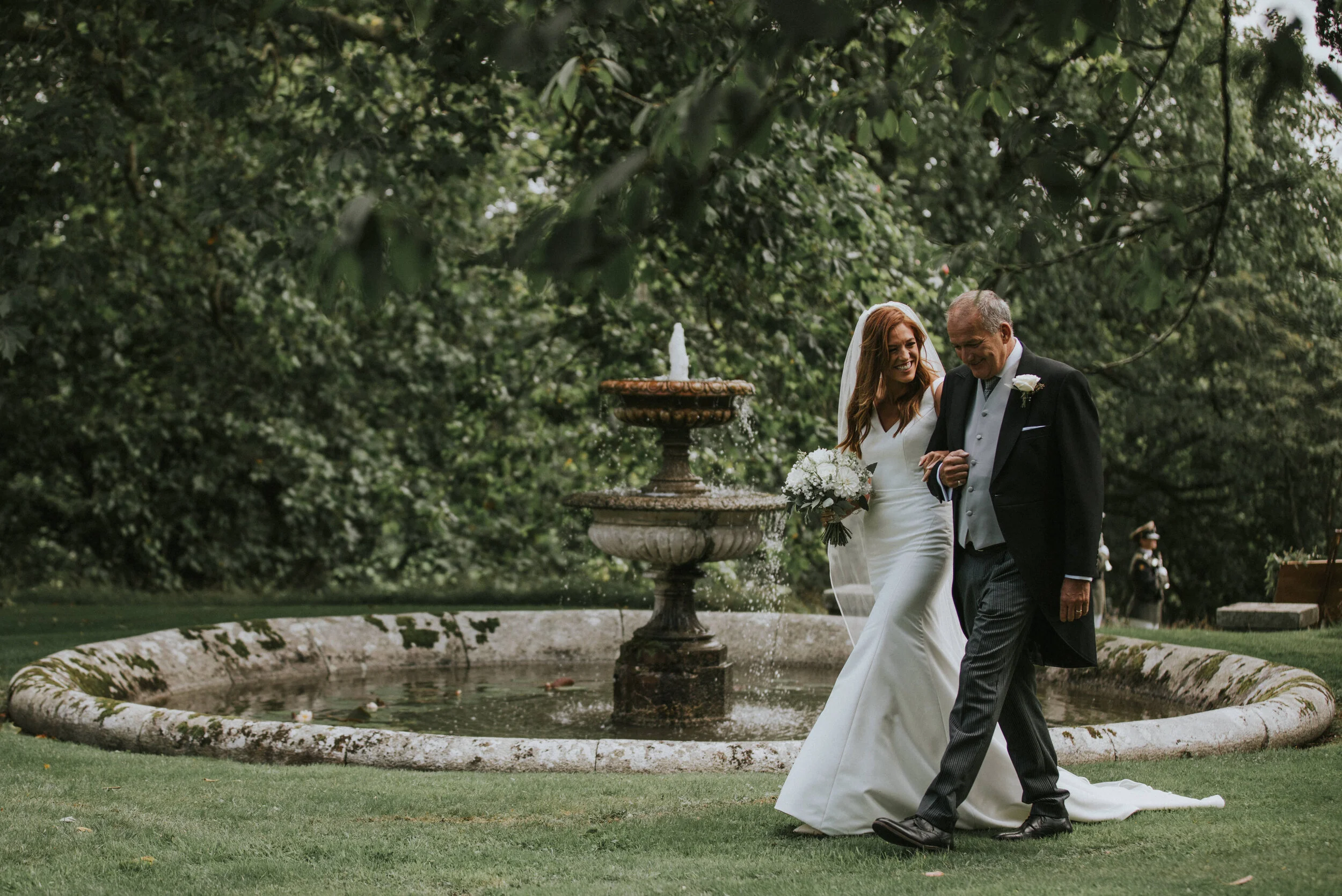 A bride in a white wedding dress holding a bouquet walks beside an older man in a tuxedo near a fountain in a lush green garden.