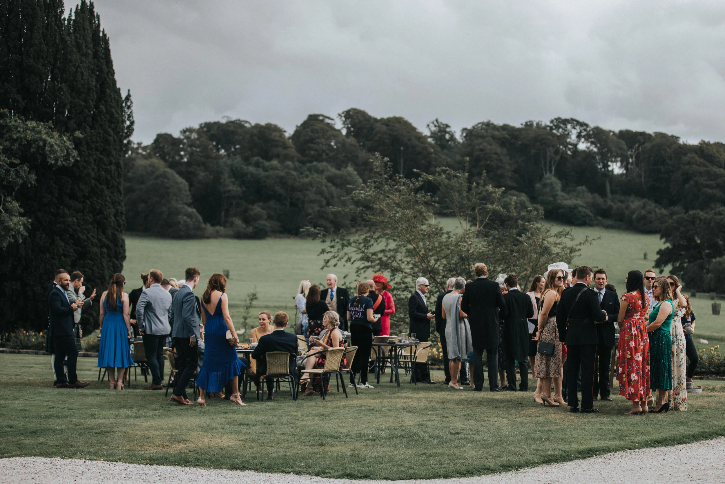 A large outdoor gathering of people dressed in formal and semi-formal attire, standing and sitting on a lawn with tables, against a backdrop of green fields and trees under cloudy sky.