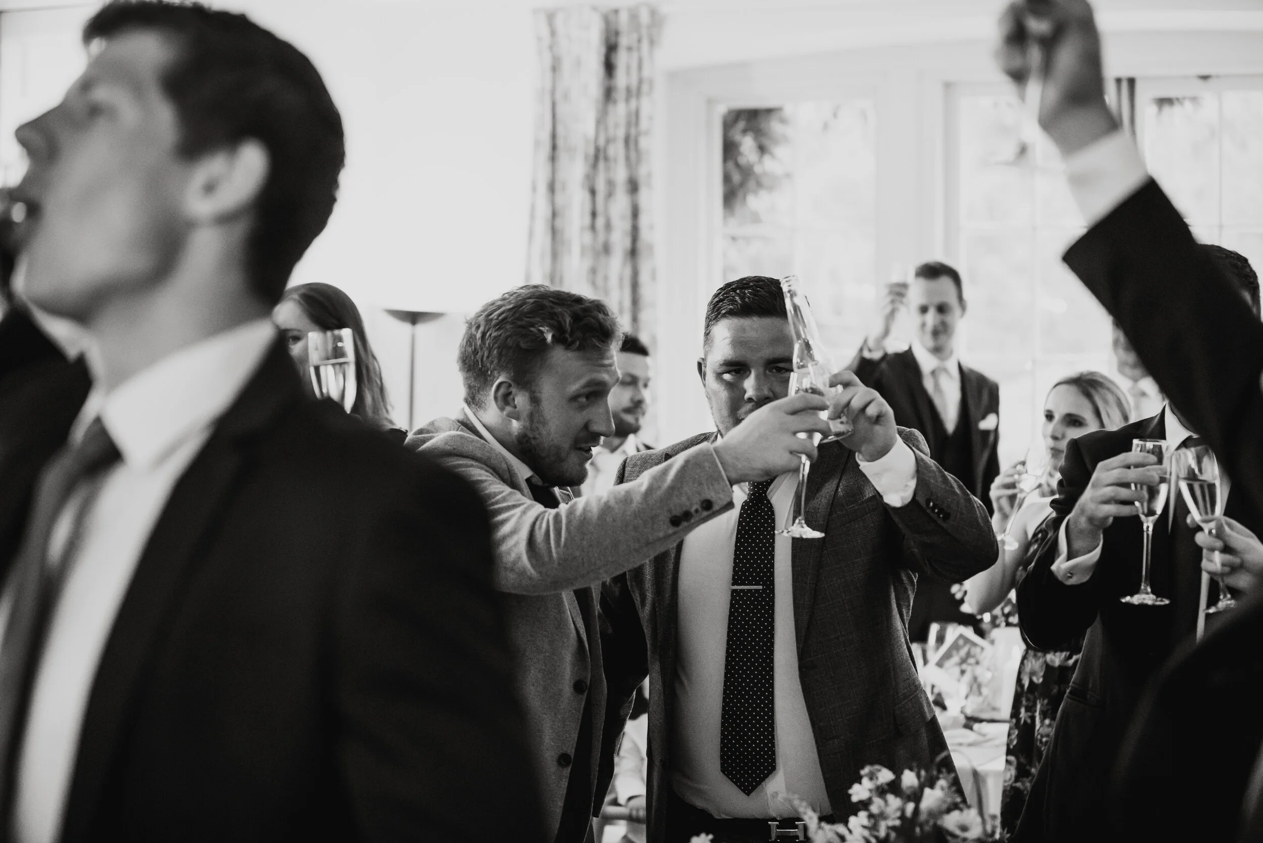 People at a formal gathering or celebration, raising glasses in a toast. Men in suits, women in elegant attire, in a bright room with large windows.