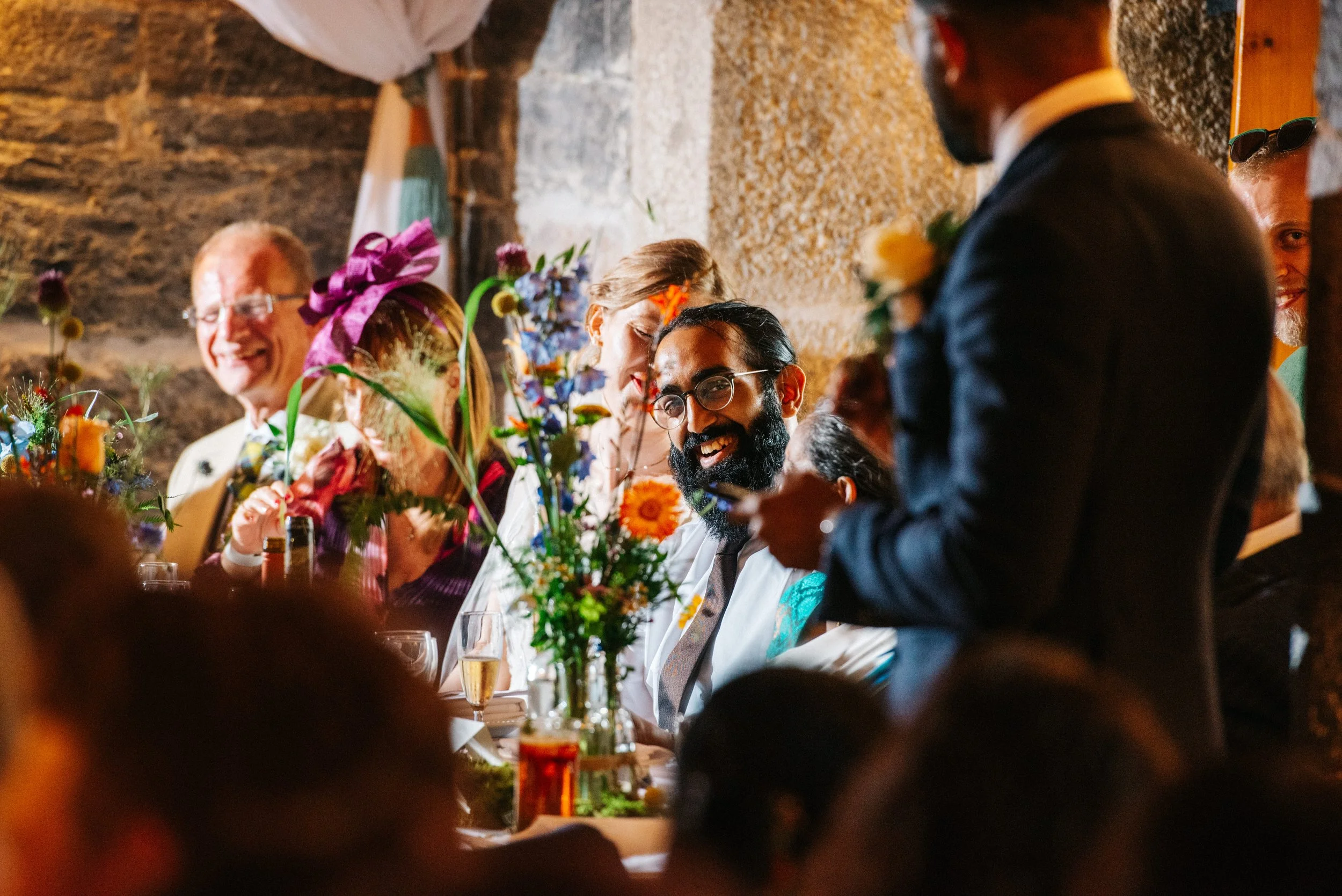 People sitting at a reception table during a celebration, with colorful flowers and glasses of sparkling wine. The focus is on a smiling man with a beard and glasses, wearing a white shirt, and a man standing and speaking, dressed in a dark suit with