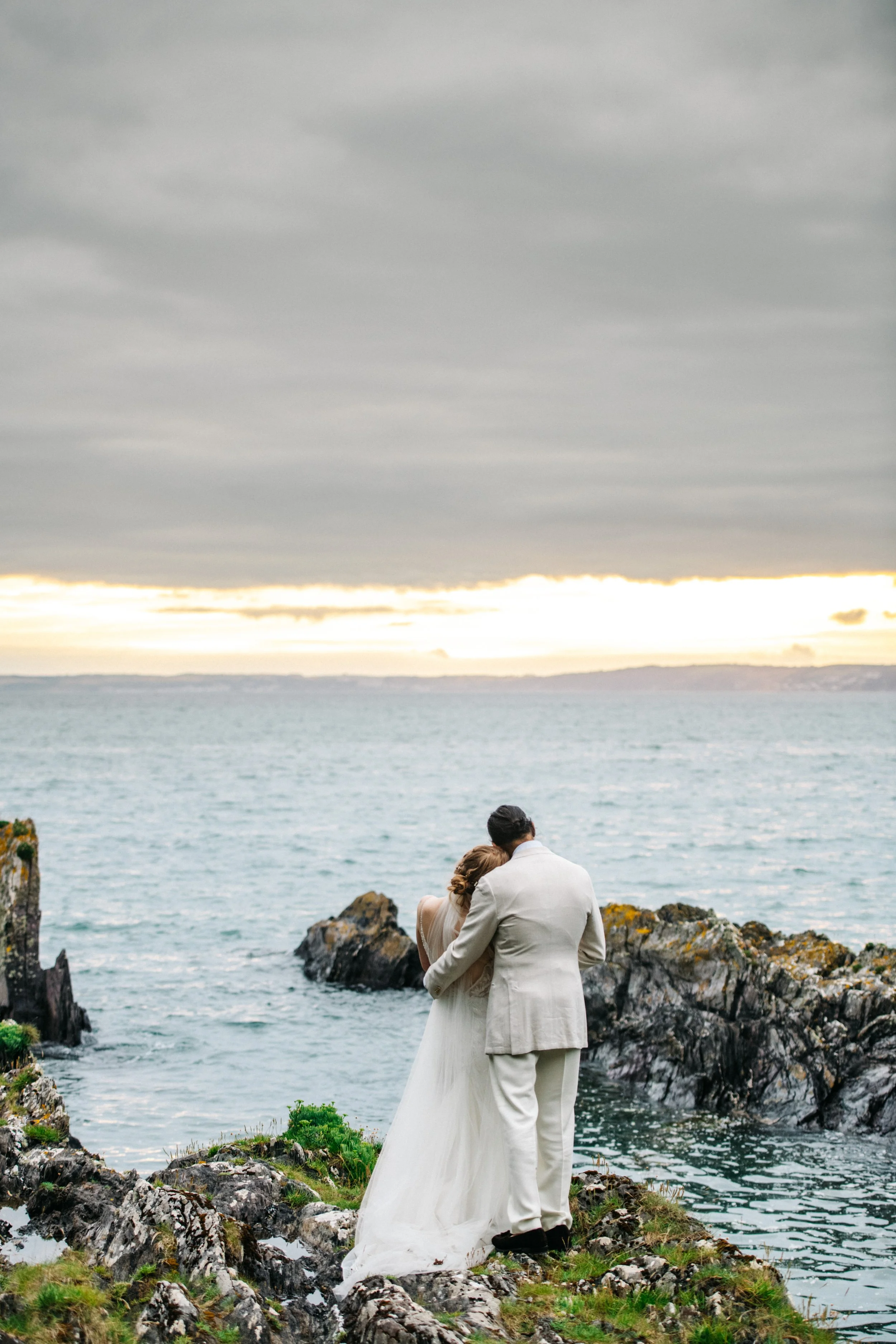 A bride and groom standing on rocky land by the sea, embracing each other at sunset under cloudy skies.