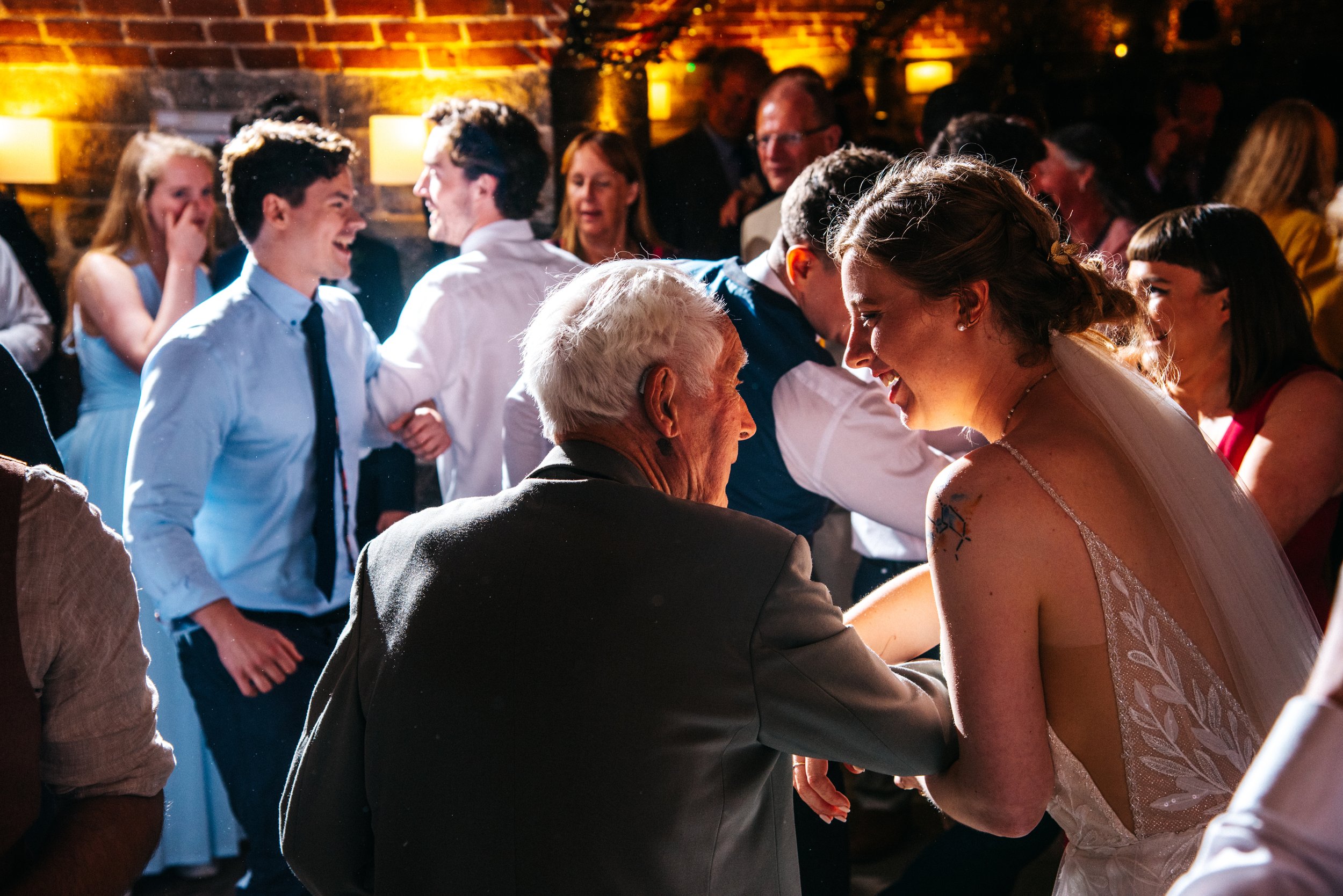 People dancing and celebrating at a wedding reception, with a bride and an elderly man in focus, smiling and enjoying the moment.