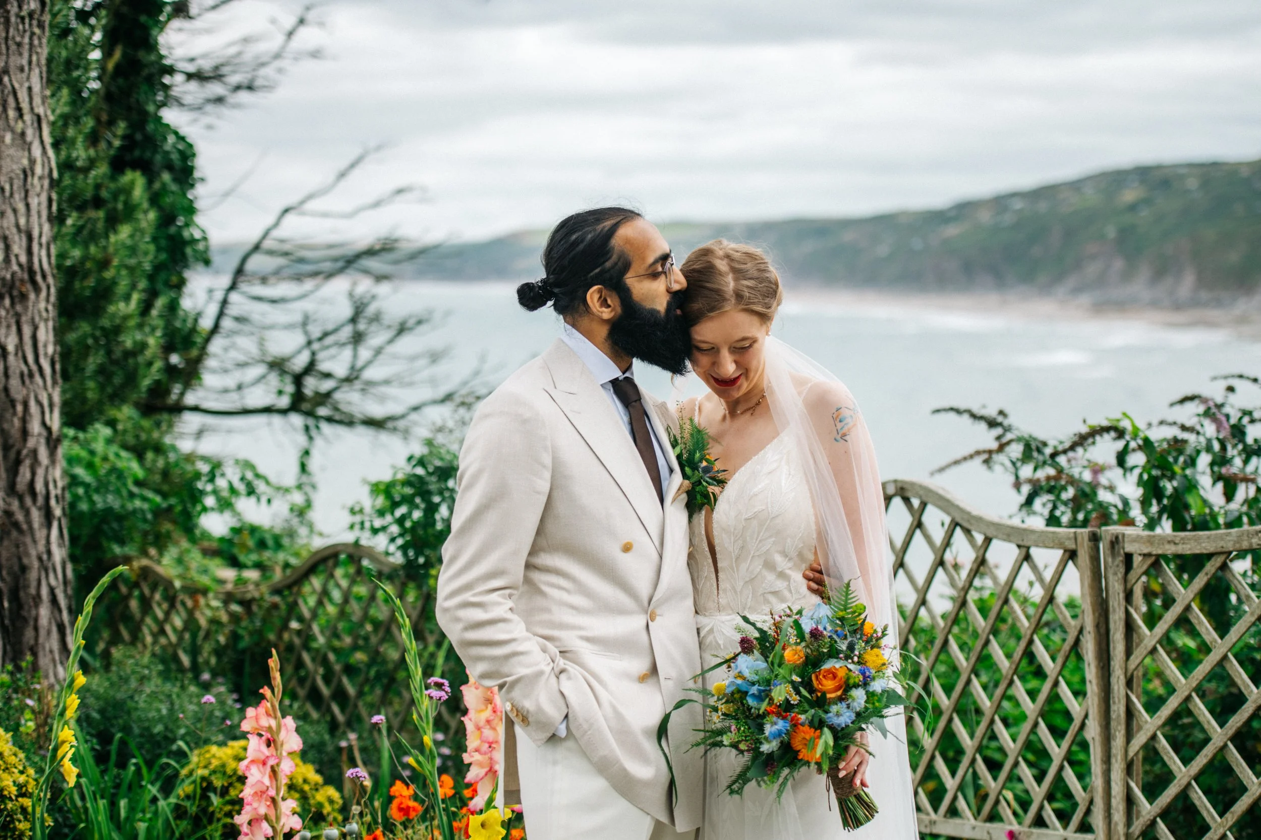 A newlywed couple stands outdoors by a garden fence overlooking the ocean. The groom, with dark hair and beard, wearing a white suit with a boutonniere, kisses the bride on the forehead. The bride, with light brown hair and a tattoo on her shoulder, 