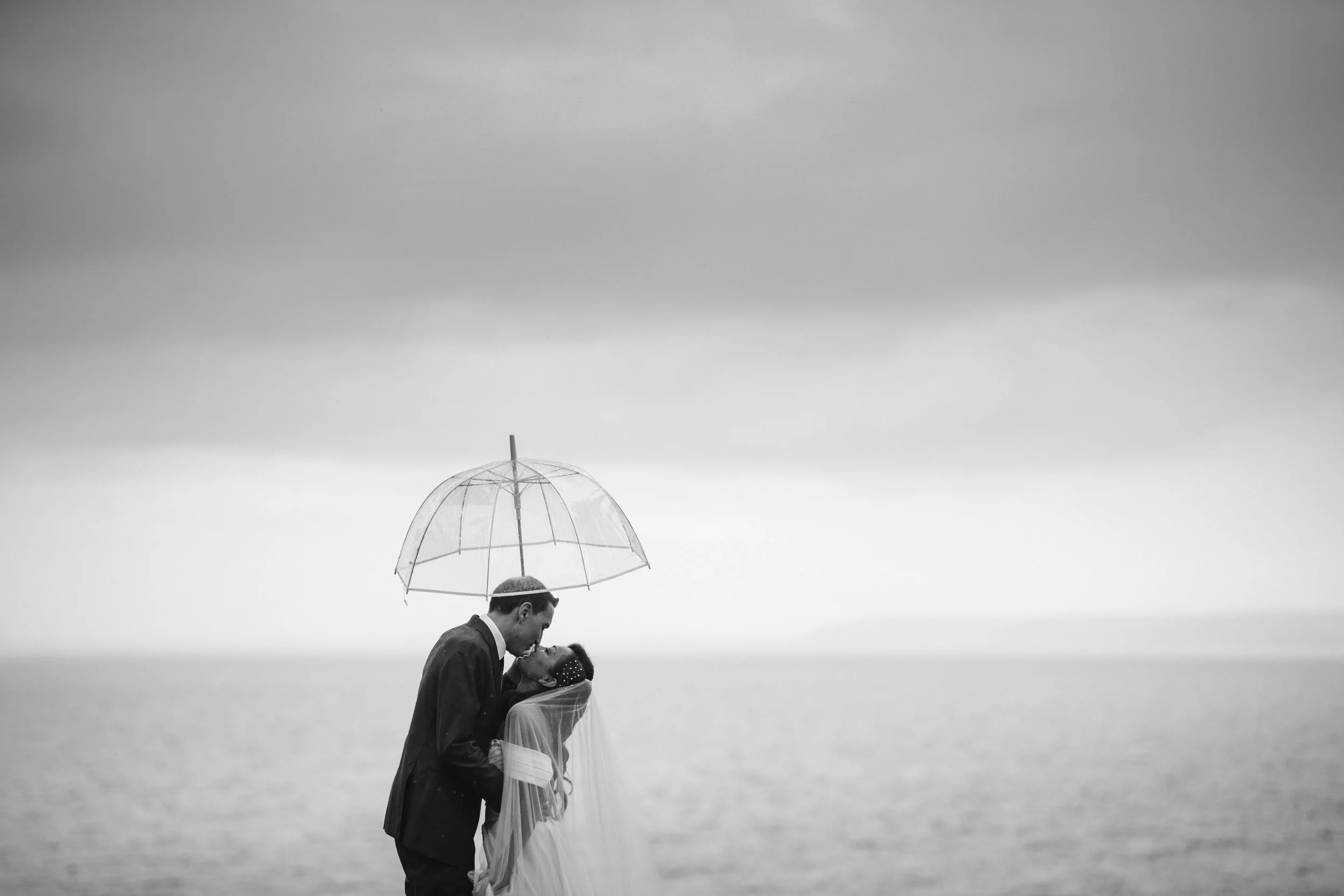 A black-and-white photo of a couple in wedding attire sharing a kiss under an umbrella near the water on a cloudy day.