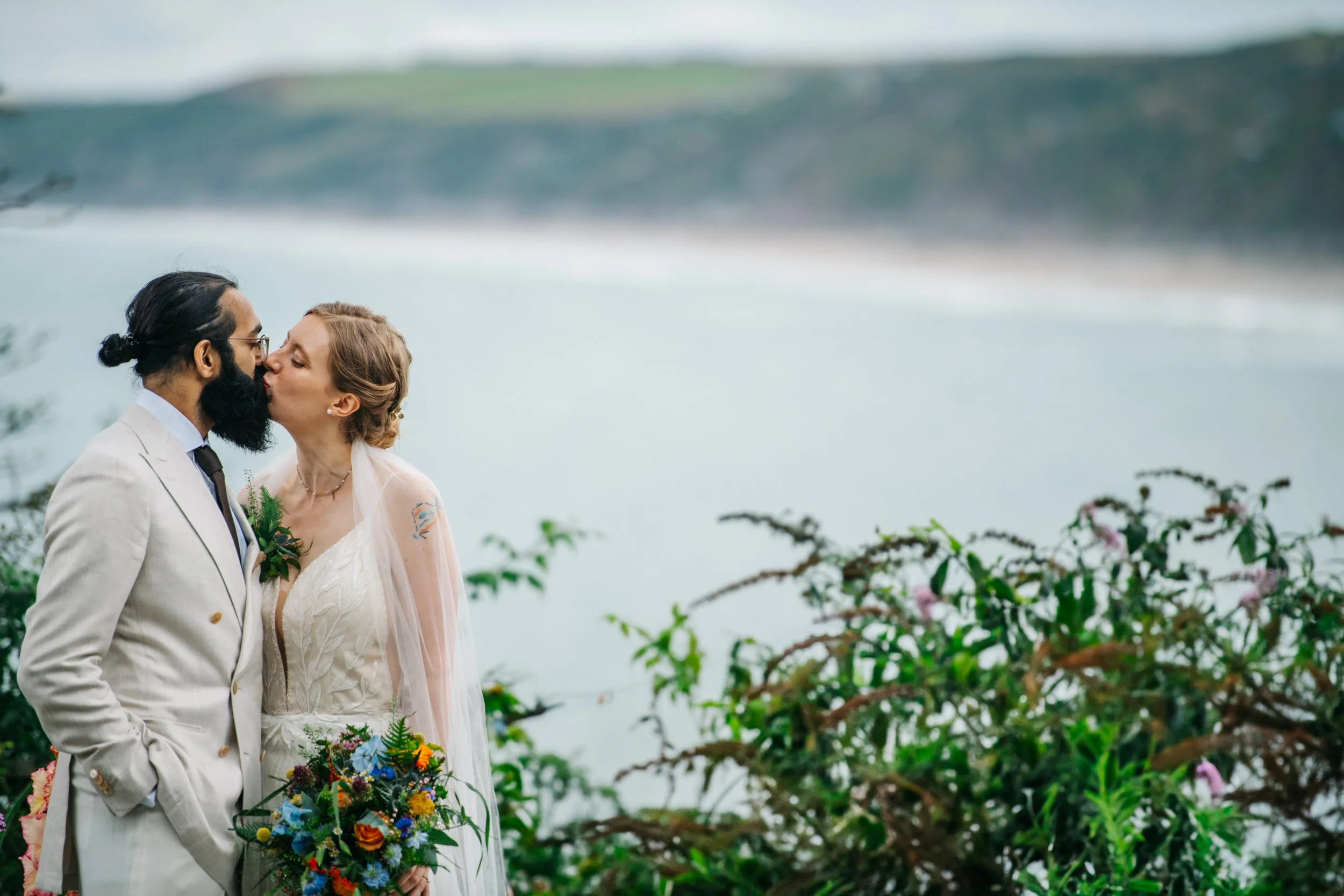 A bride and groom kissing outdoors near a body of water with lush greenery and flowering plants in the foreground.