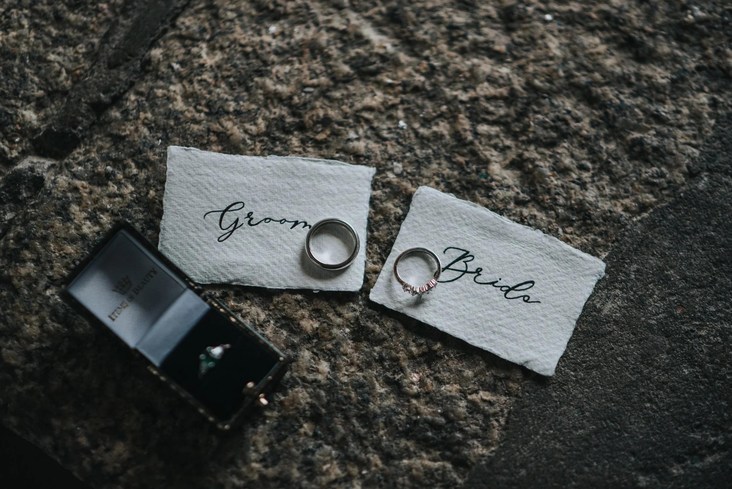 Wedding rings on paper labels labeled "Groom" and "Bride" on a rough stone surface, with an open jewelry box nearby.