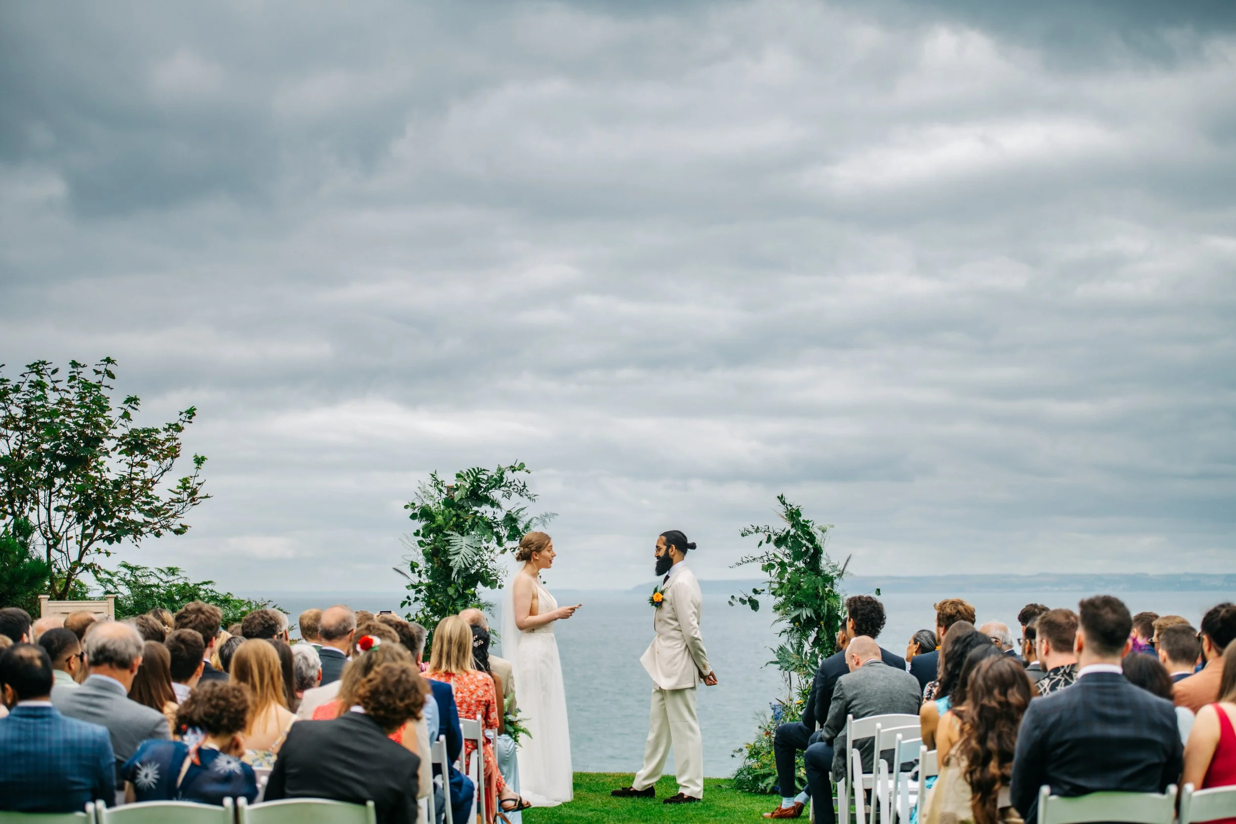 A wedding ceremony taking place outdoors near the water under cloudy skies, with the couple standing at the front facing each other, surrounded by guests seated on white chairs.