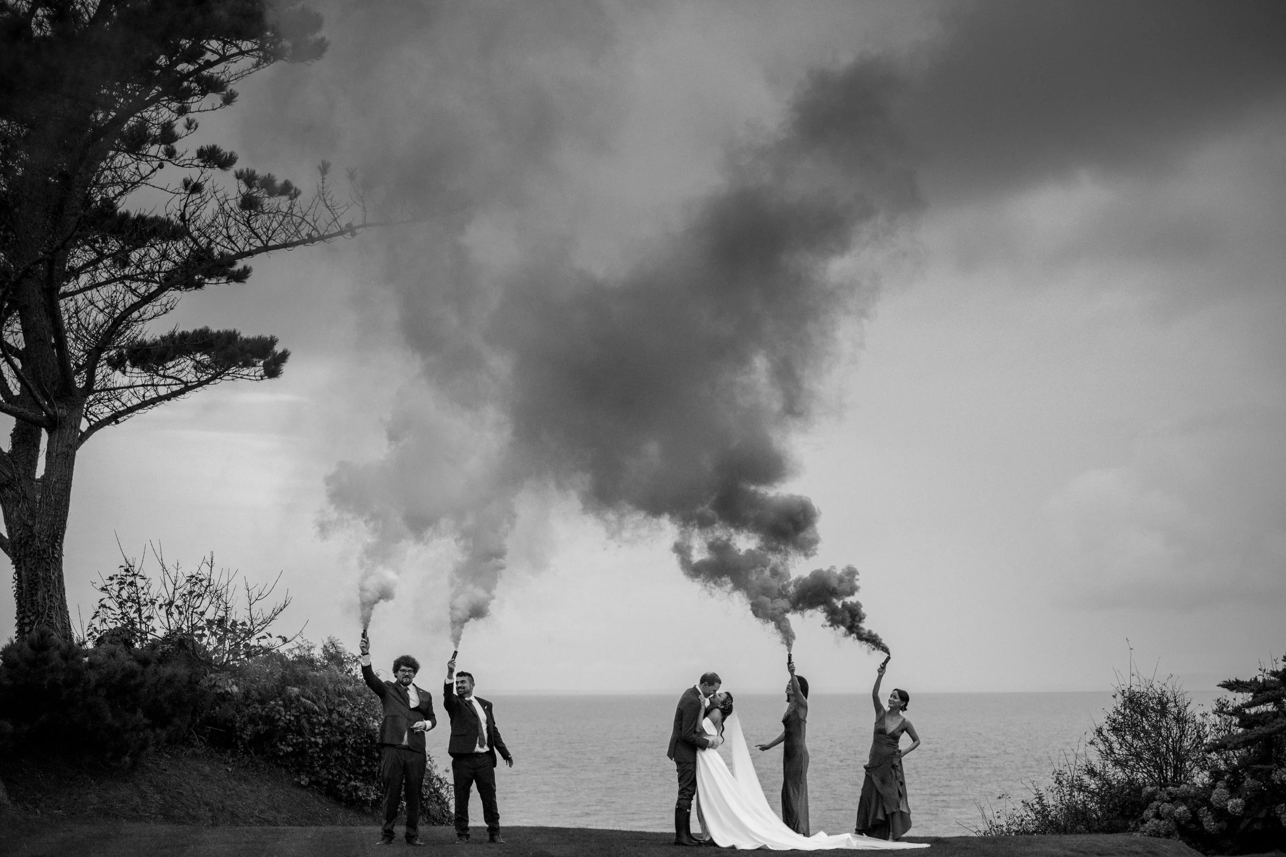 A wedding photo set outdoors by the ocean in black and white. Three men are holding smoke bombs emitting dark smoke, and a bride and groom are kissing in front of them. The bride wears a long white dress, and the sculpted groom is in a suit. Two wome