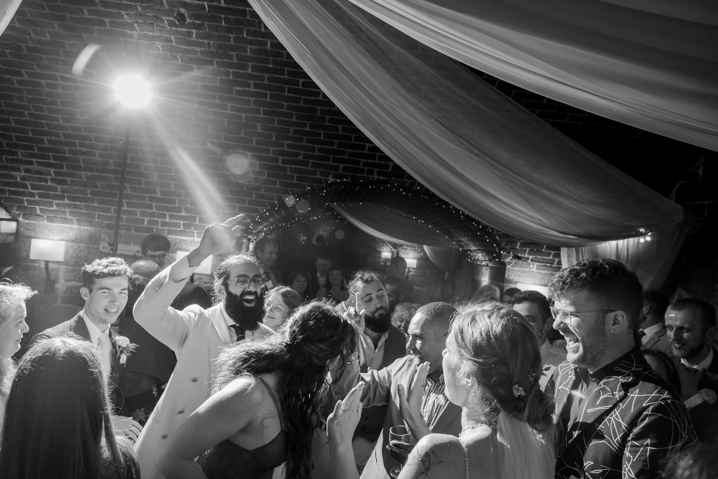 Group of people celebrating and dancing at a wedding reception in a decorated venue with brick walls and draped fabric ceiling.