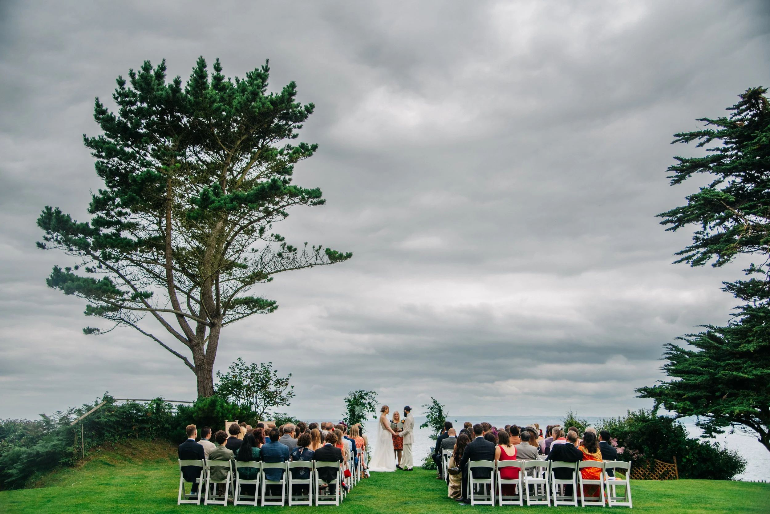 A wedding ceremony outdoors with a couple exchanging vows, seated guests on both sides, under a large tree, with dark cloudy sky overhead.