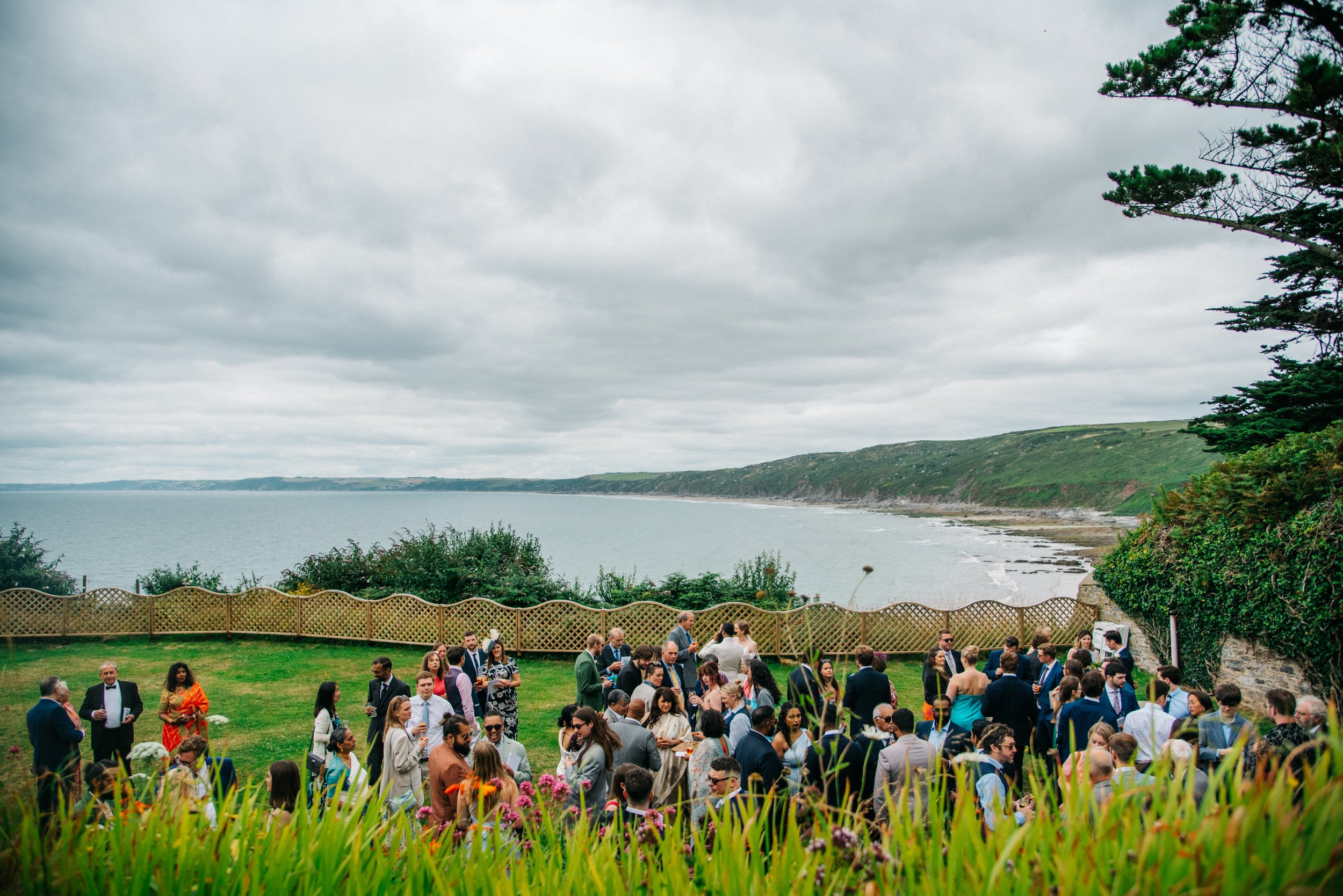 Group of people gathered outdoors near a coastline with green hills and cloudy sky in the background, likely at a wedding reception.
