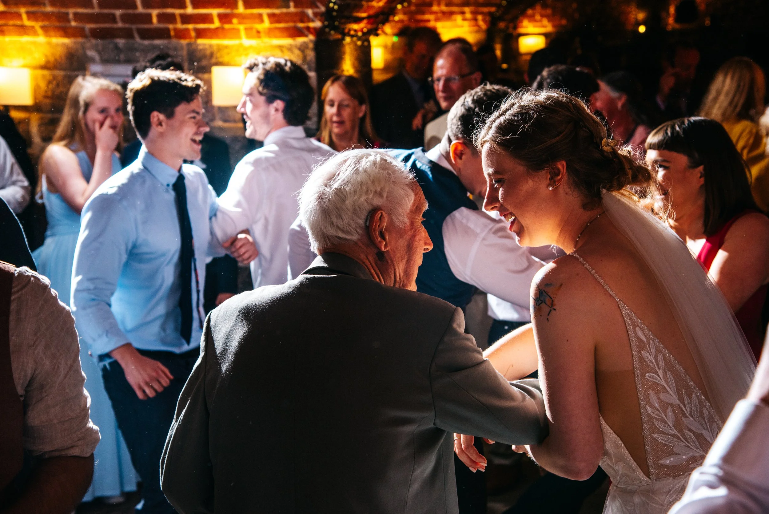 A wedding reception scene with a bride dancing and smiling with an elderly man, surrounded by guests dancing and celebrating in a warmly lit venue with exposed brick walls.
