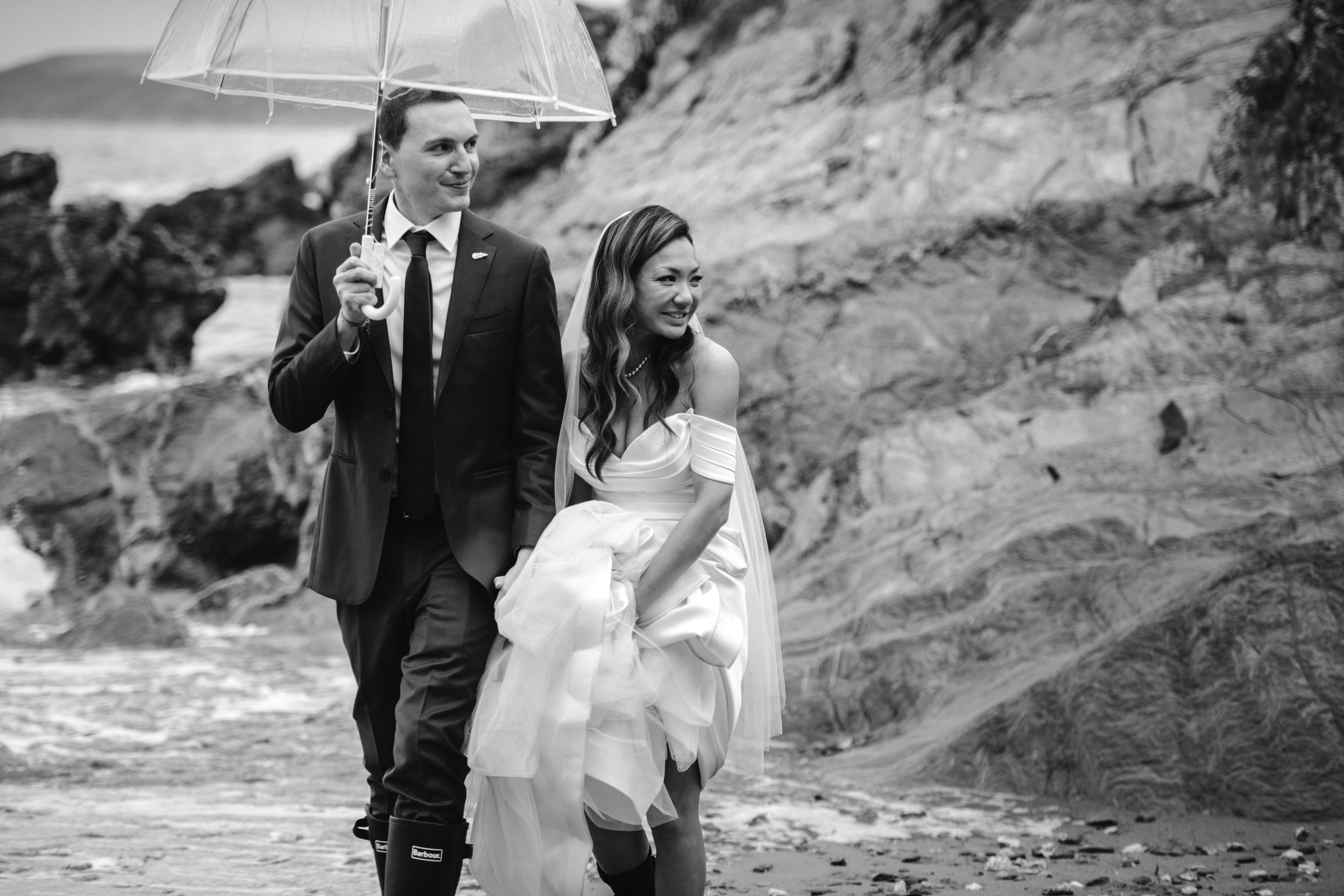 A bride and groom walking on a rocky beach, with the groom holding an umbrella over both of them. The bride is smiling and holding up her wedding dress.