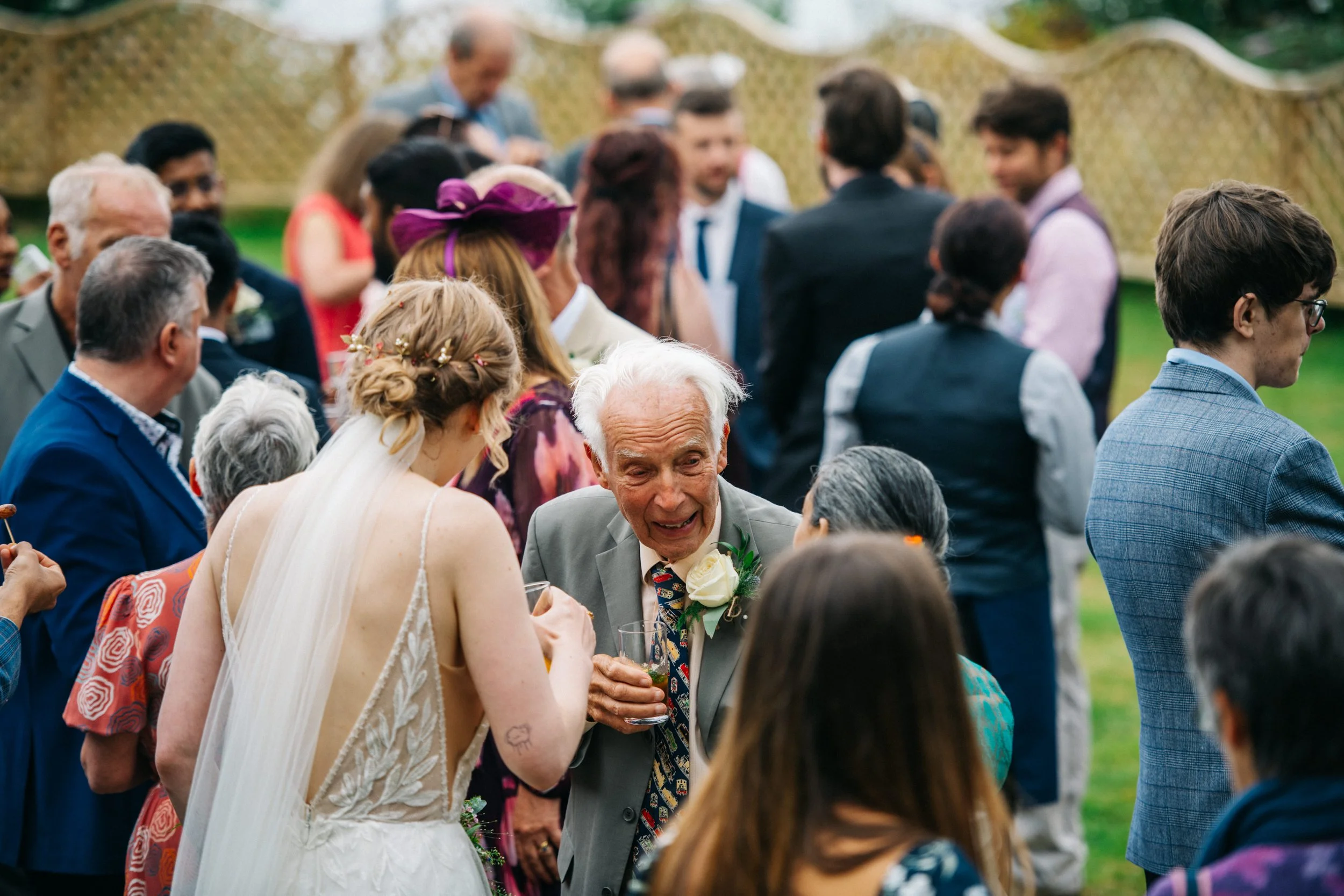 At a wedding reception outdoors, a young bride in a white dress with a veil and floral hairpiece is talking with an elderly man in a gray suit holding a glass.