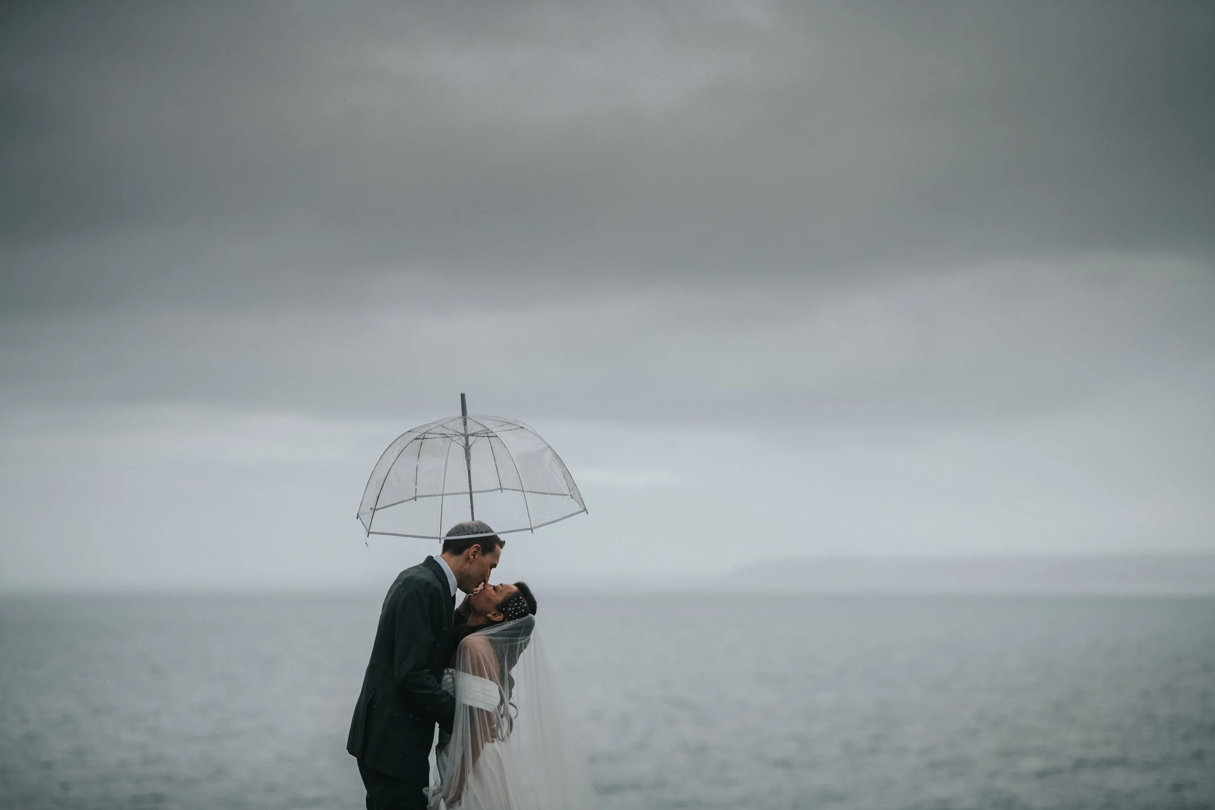 A couple in wedding attire kissing under a transparent umbrella on a cloudy, rainy day by the water.