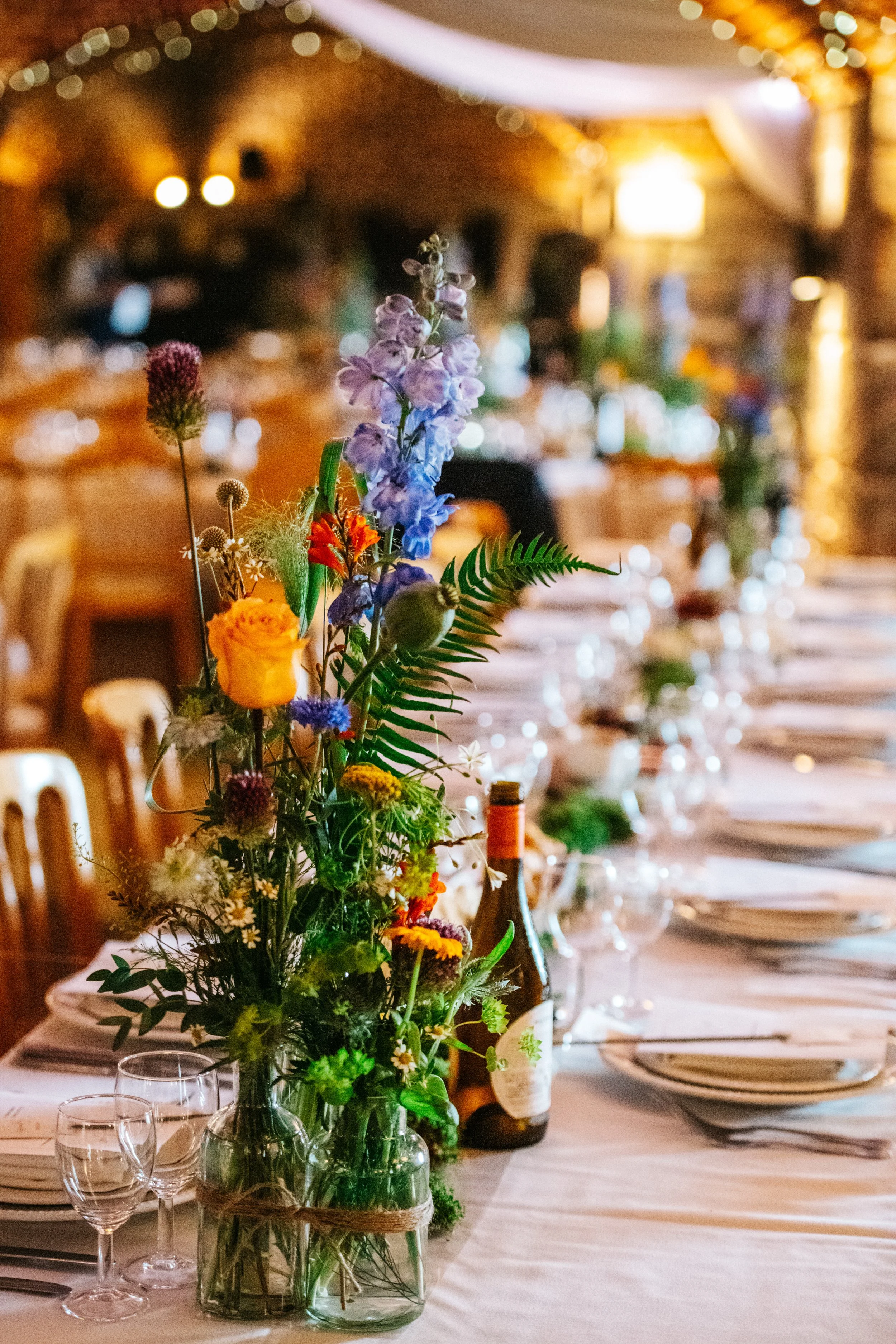 A decorated dining table with floral centerpiece, glassware, plates, and bottles, set in a warmly lit venue for an event.