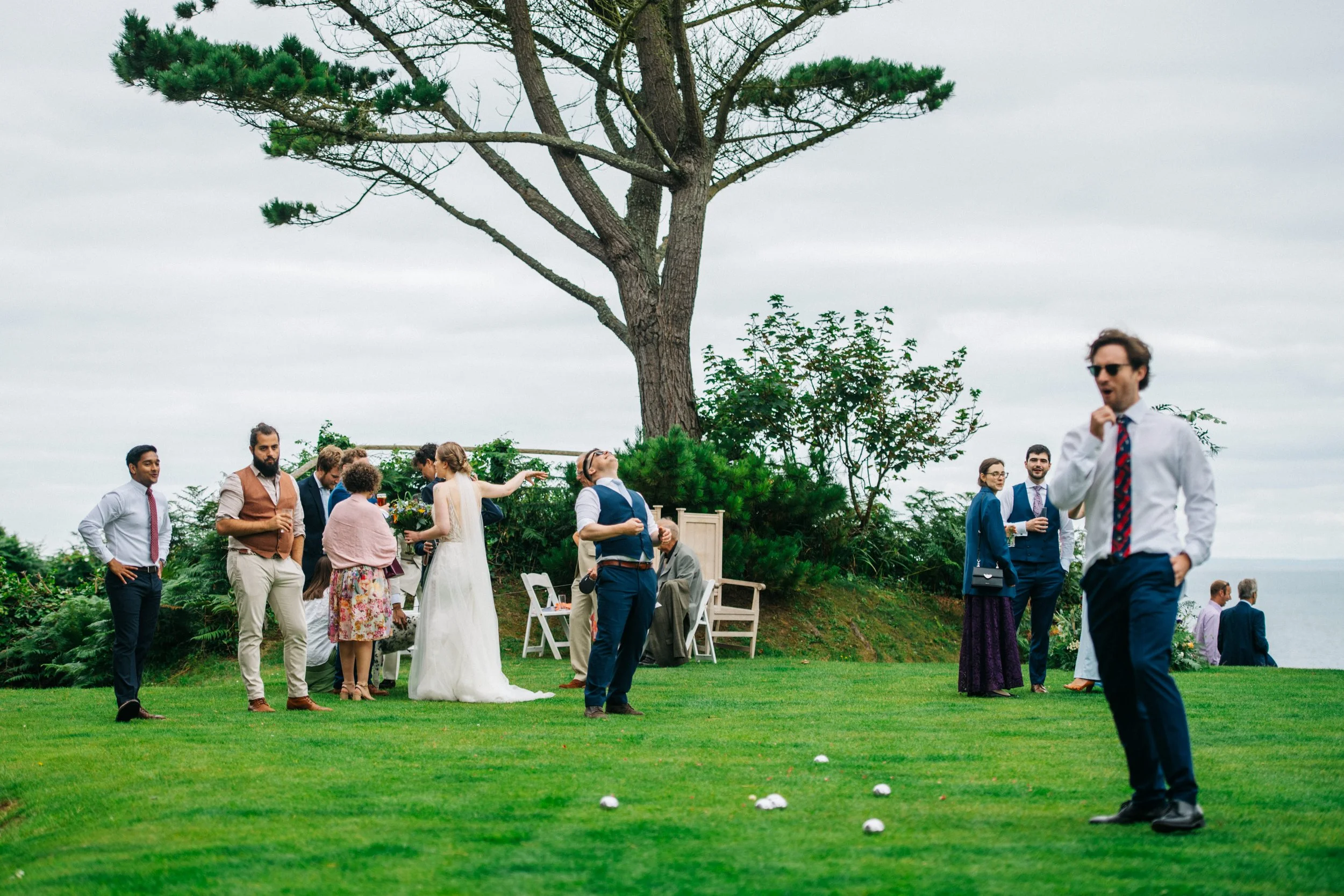 People attending a wedding reception outdoors on a grassy area with a large tree, with some playing a game and others talking, under an overcast sky.