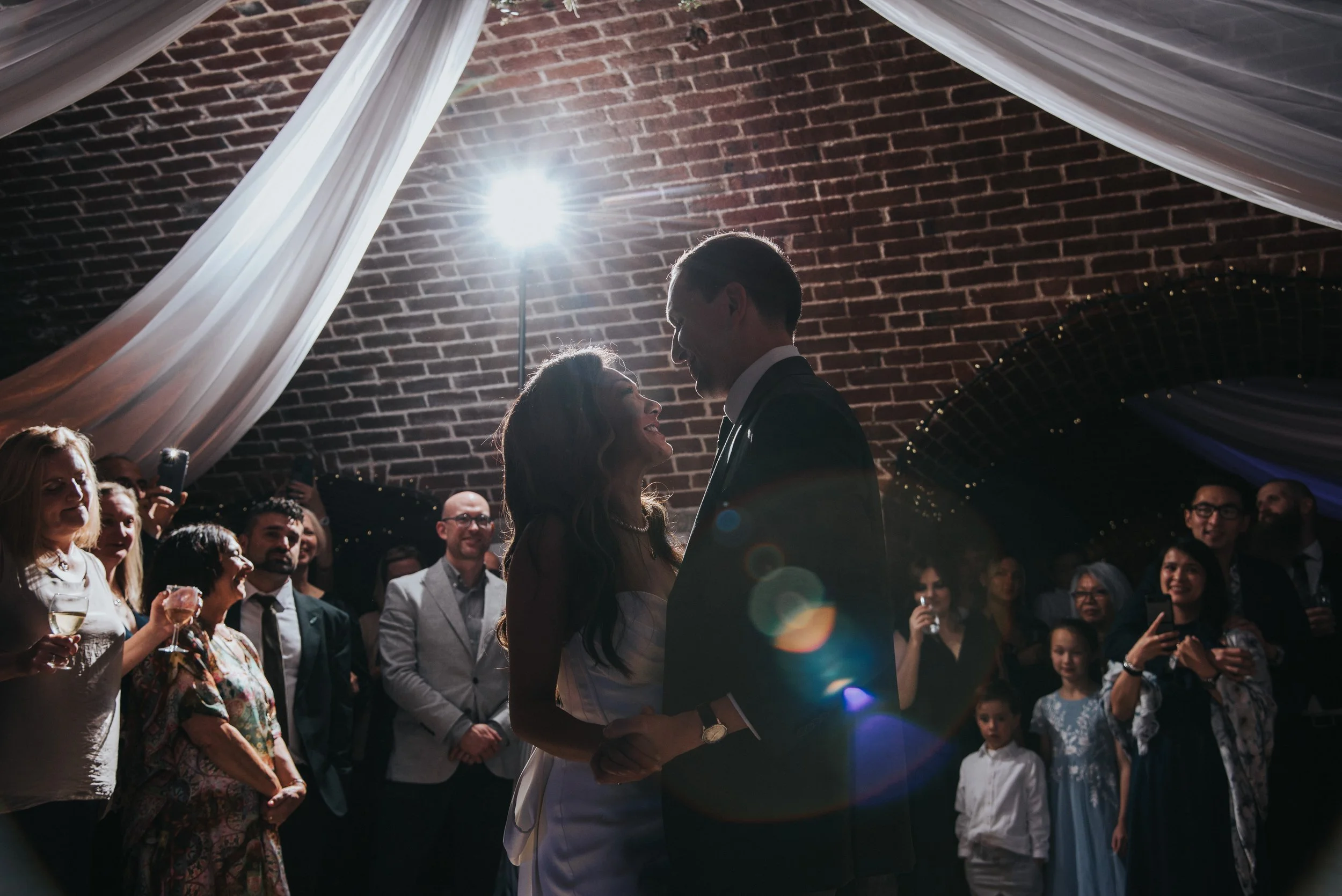 A bride and groom share a dance at their wedding reception under a decorated ceiling with white drapes and brick arch, surrounded by guests with drinks, some taking photos, and smiling.