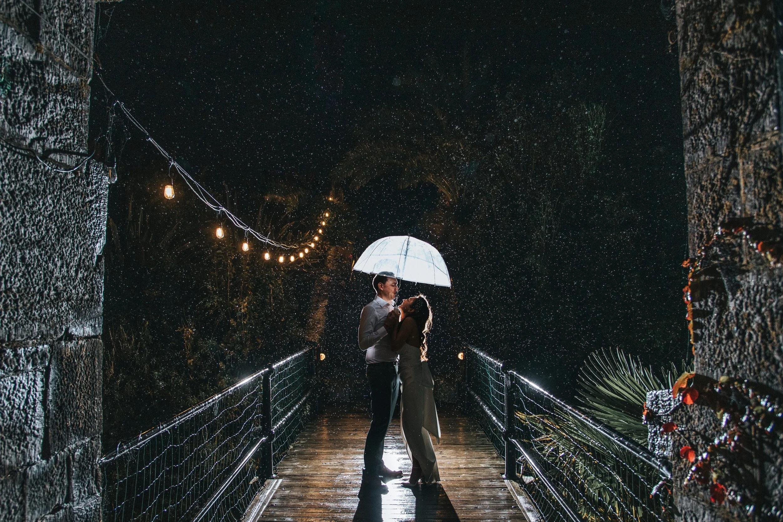 A couple stands on a wooden bridge at night, sharing an umbrella in the rain. They are close, looking into each other's eyes, with lights strung overhead and raindrops falling around them.
