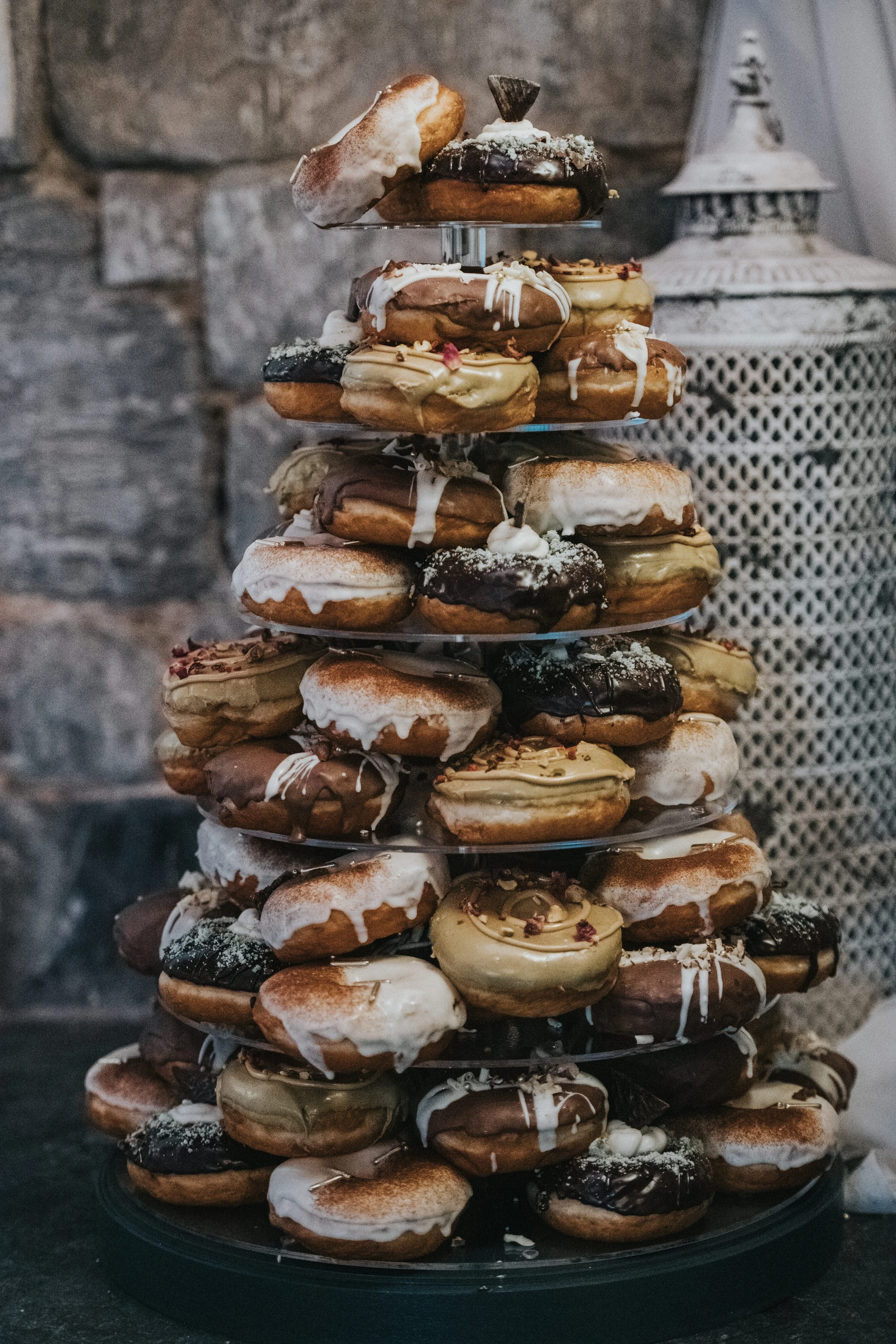 A multi-tiered stand filled with assorted donuts covered in chocolate, icing, and sprinkles, set against a rustic stone wall background.