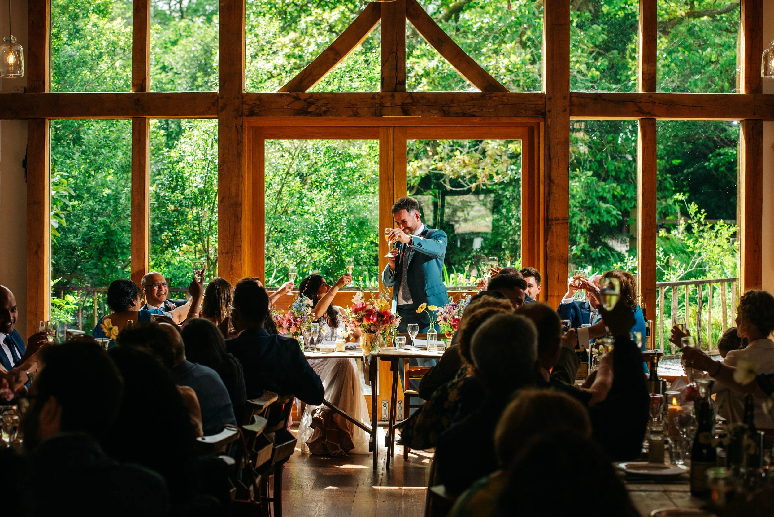 A man in a blue suit giving a toast at a wedding reception, surrounded by seated guests raising glasses, with a large window and greenery outside