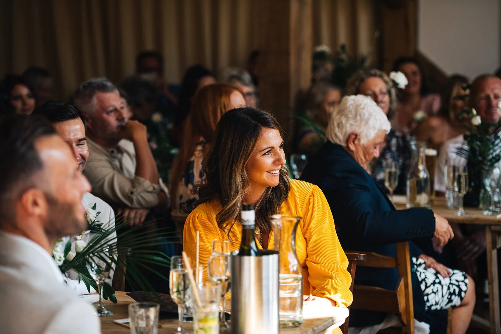 People sitting at long table during a celebration or event, smiling and enjoying the occasion, with drinks and decorative elements on the table.