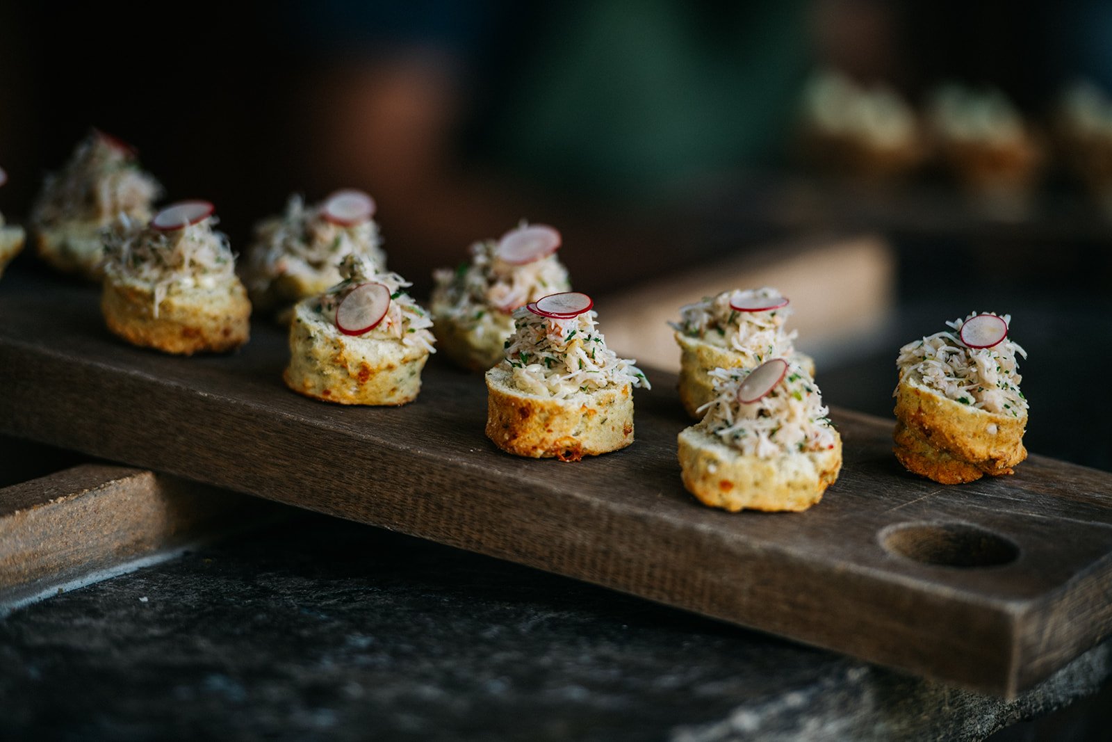 Small appetizers topped with shredded crab meat, thin radish slices, and chopped herbs on a wooden serving board.
