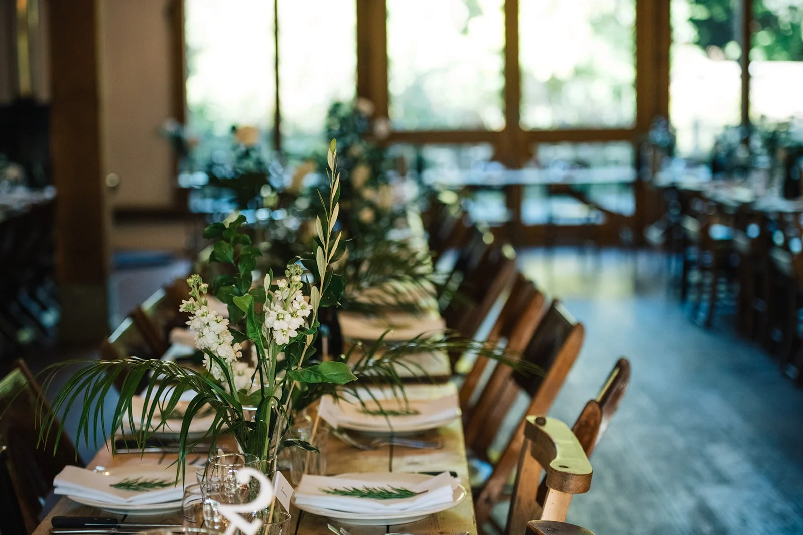 A long dining table set for a formal event, decorated with a floral centerpiece featuring white flowers and green foliage, in a room with large windows and wooden chairs.
