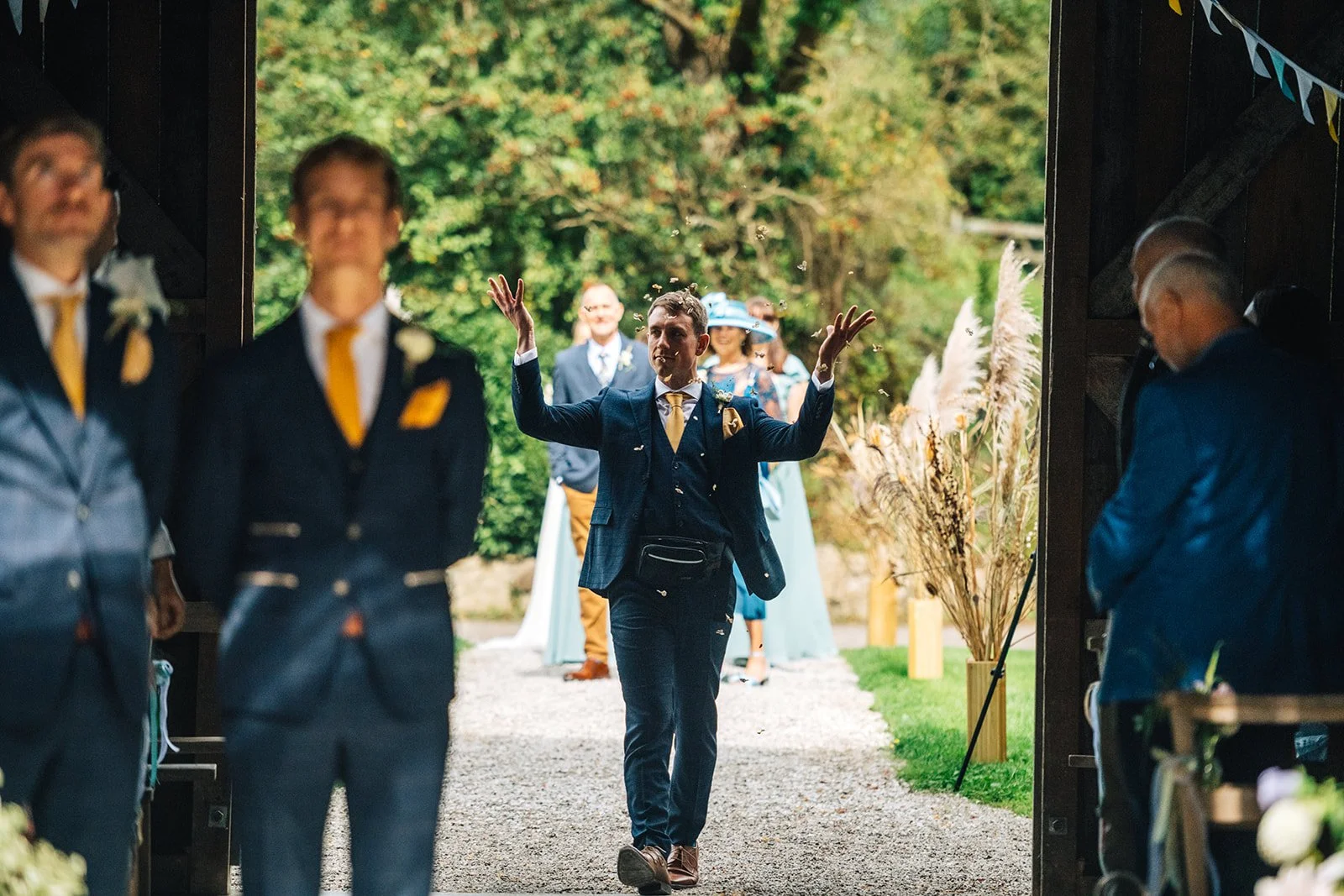 A man in a blue suit and yellow tie walks through a barn door at an outdoor wedding ceremony, with guests and wedding party members in the background.