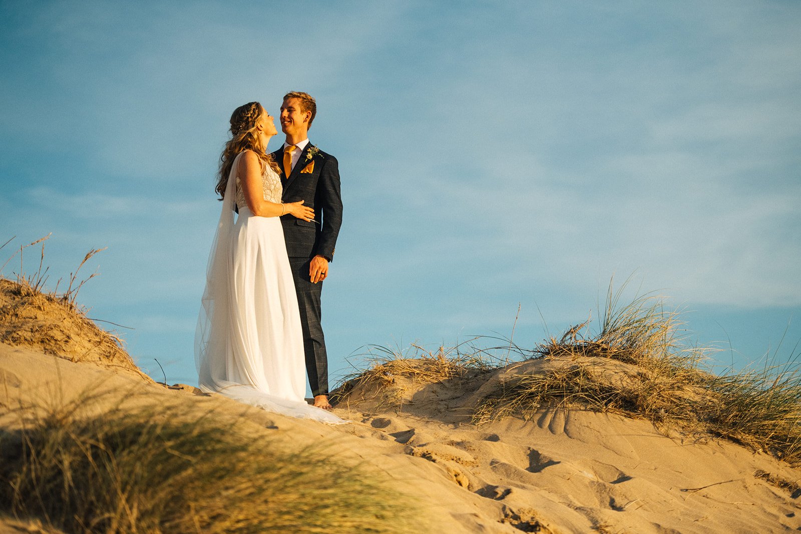 A bride and groom standing on sand dunes at the beach, sharing a joyful moment facing each other, with blue sky in the background.