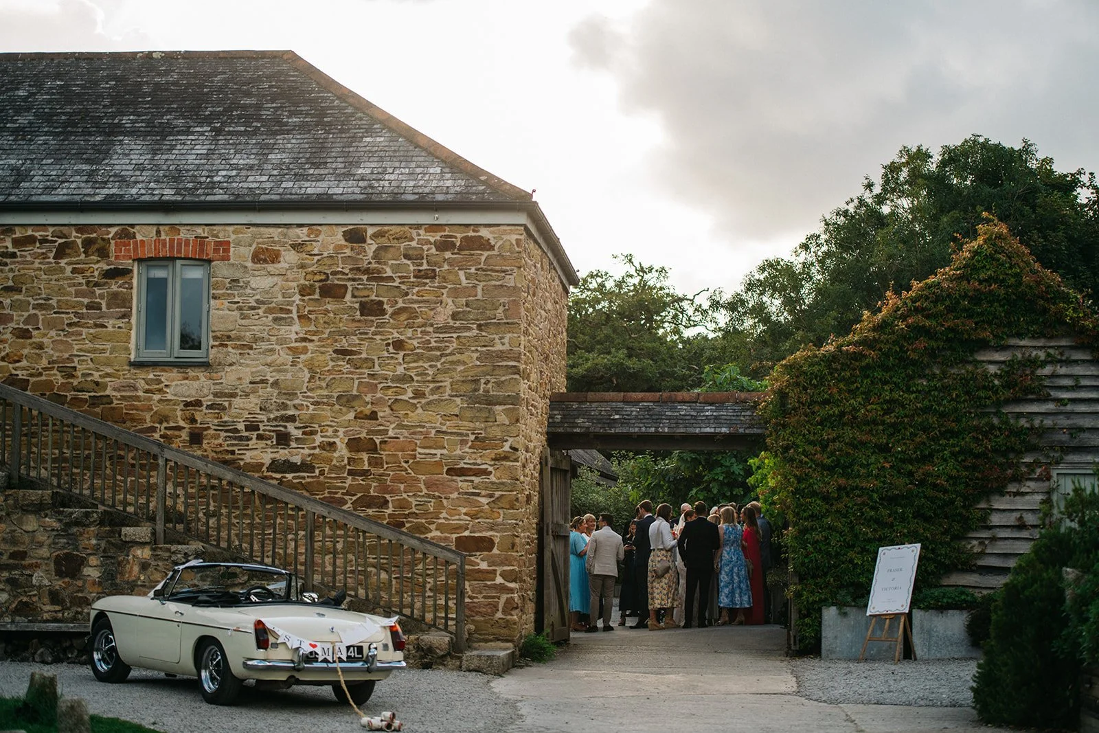 A group of people dressed in formal attire gathered outside a rustic stone building with a wooden door and a window. A vintage white convertible car with a banner reading "S ❤️ M A 4 L" is parked nearby, with a leash and dog toy on the ground. The sc