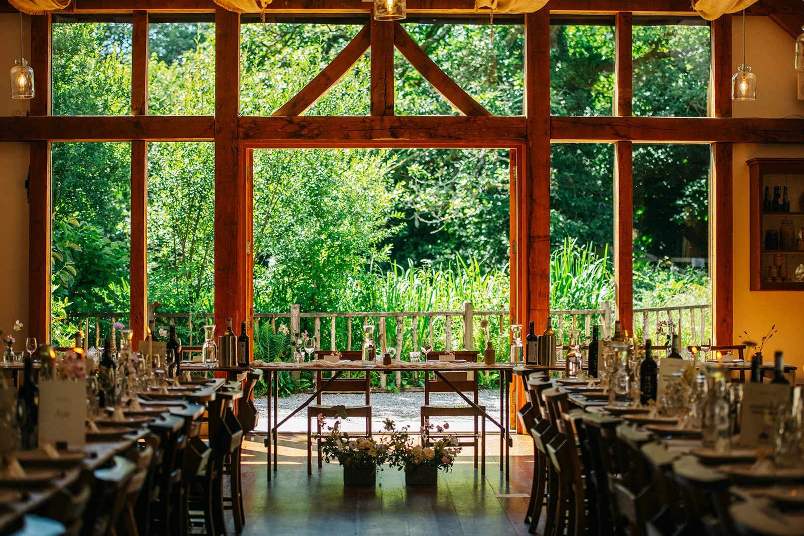 A rustic indoor dining area with wooden beams and large windows revealing a lush green outdoor garden. The tables are elegantly set with glassware, bottles, and flowers, suggesting a celebration or special event.