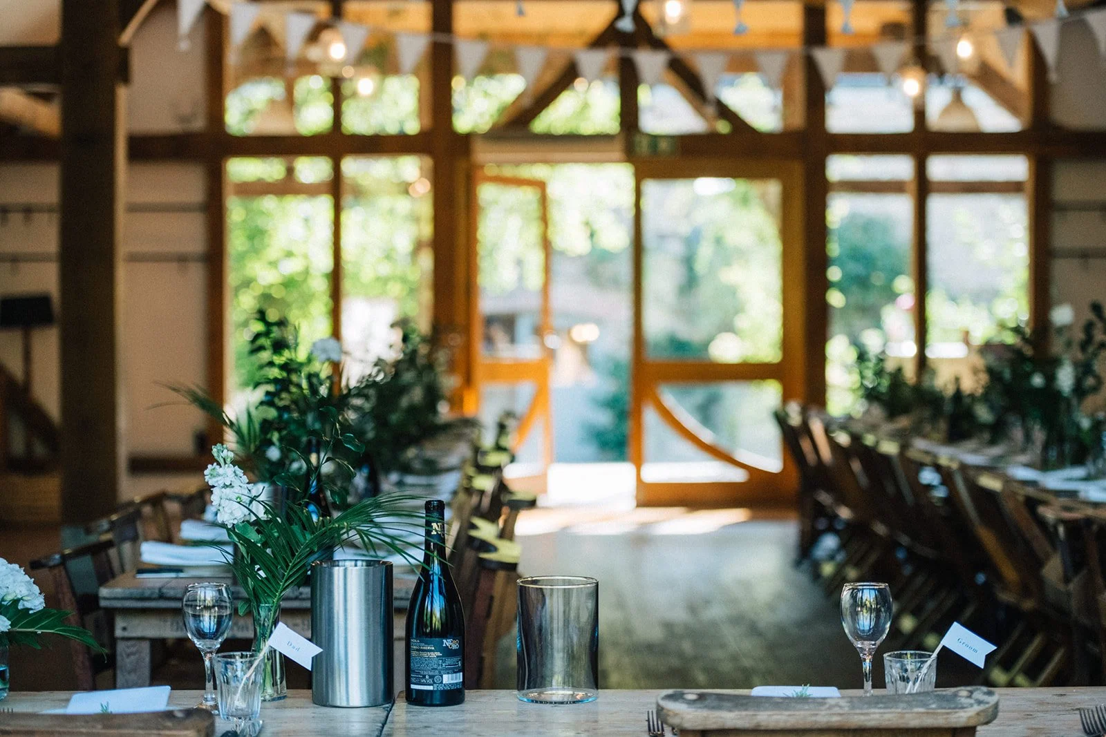 Wedding reception table set with glasses, flowers, and place cards inside a rustic wooden venue with large glass doors opening to greenery.