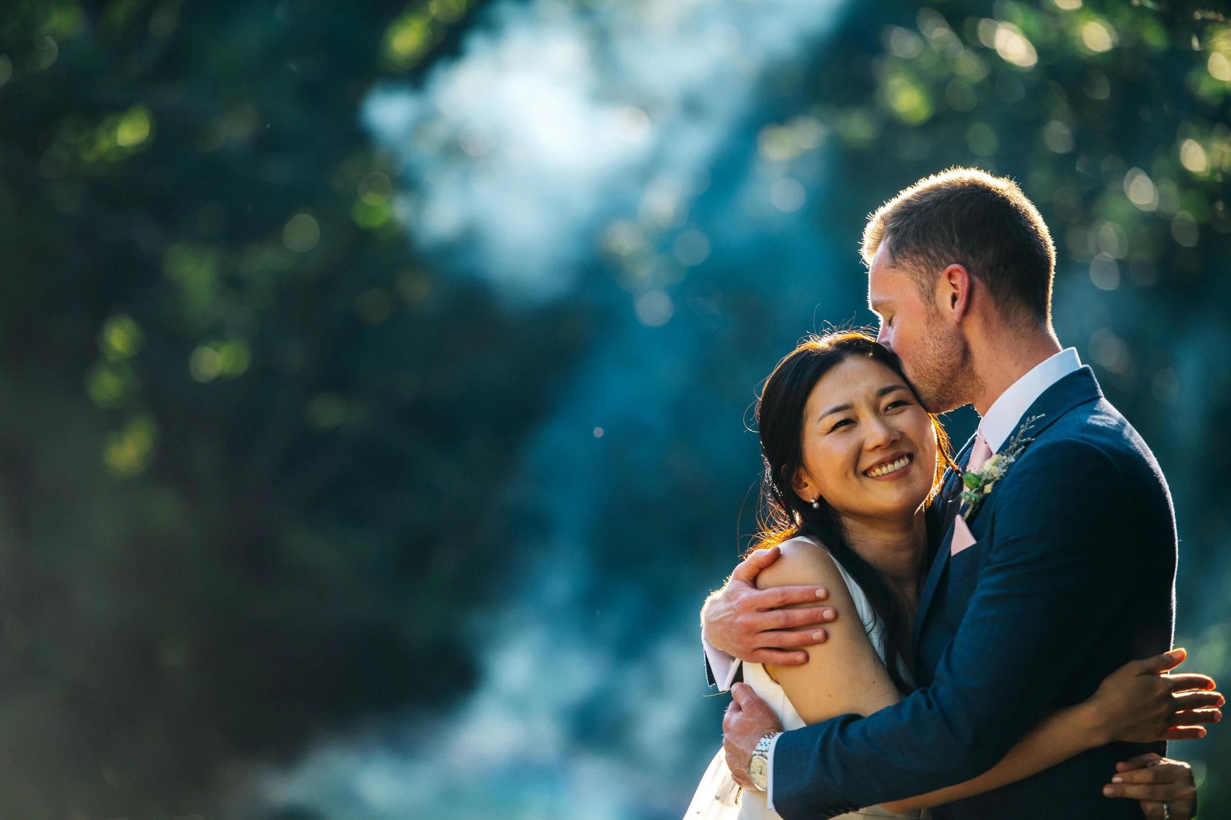A couple dressed in wedding attire embracing outdoors with trees and sunlight in the background.