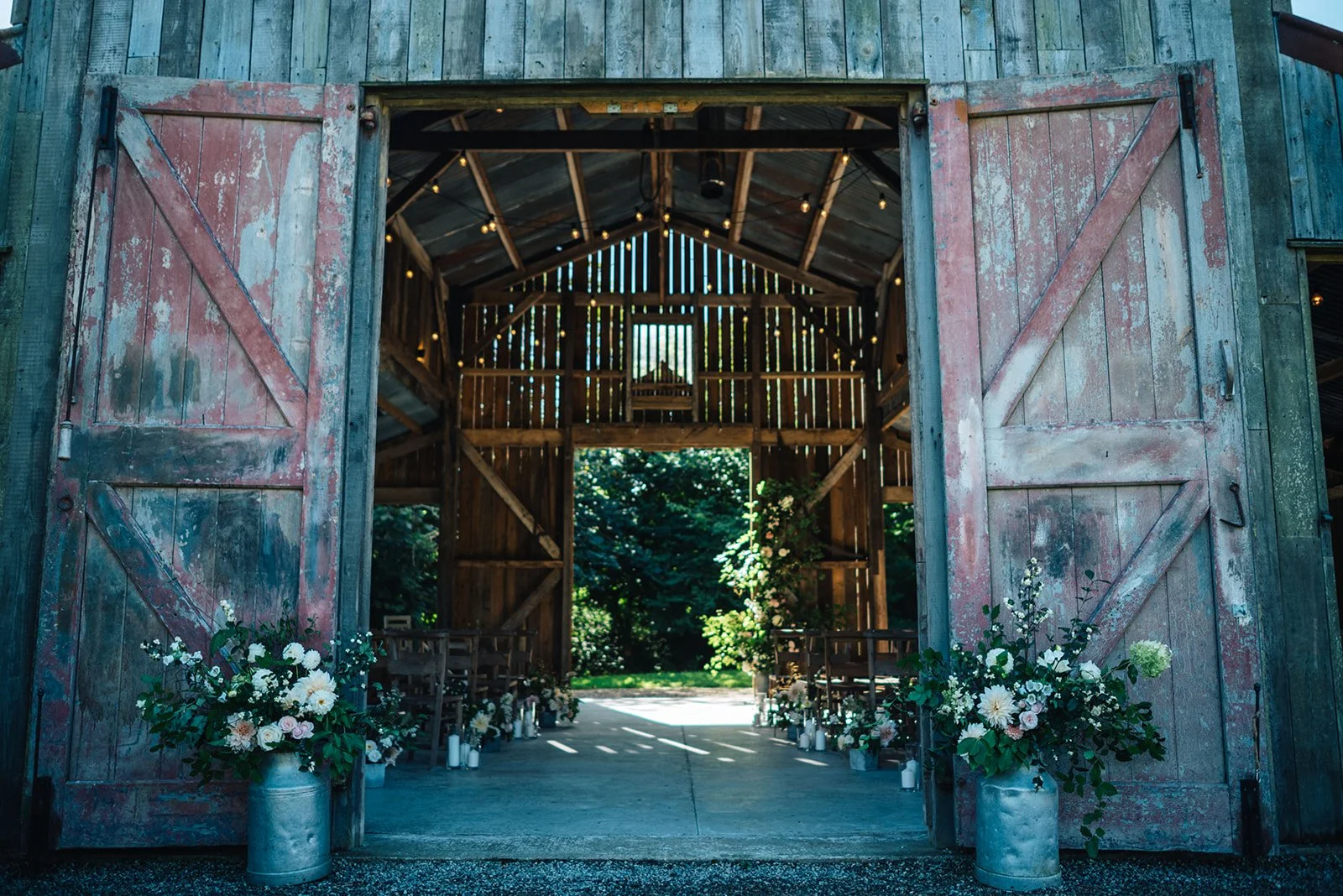 Open rustic barn doors decorated with floral arrangements and candles, revealing an outdoor wedding ceremony setup with chairs and greenery.
