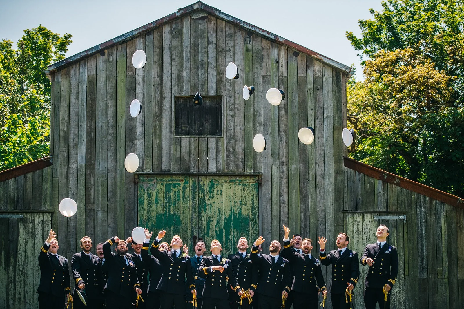 A group of men in navy blue uniforms celebrating and tossing white plates into the air outside in front of a weathered green barn with trees in the background.