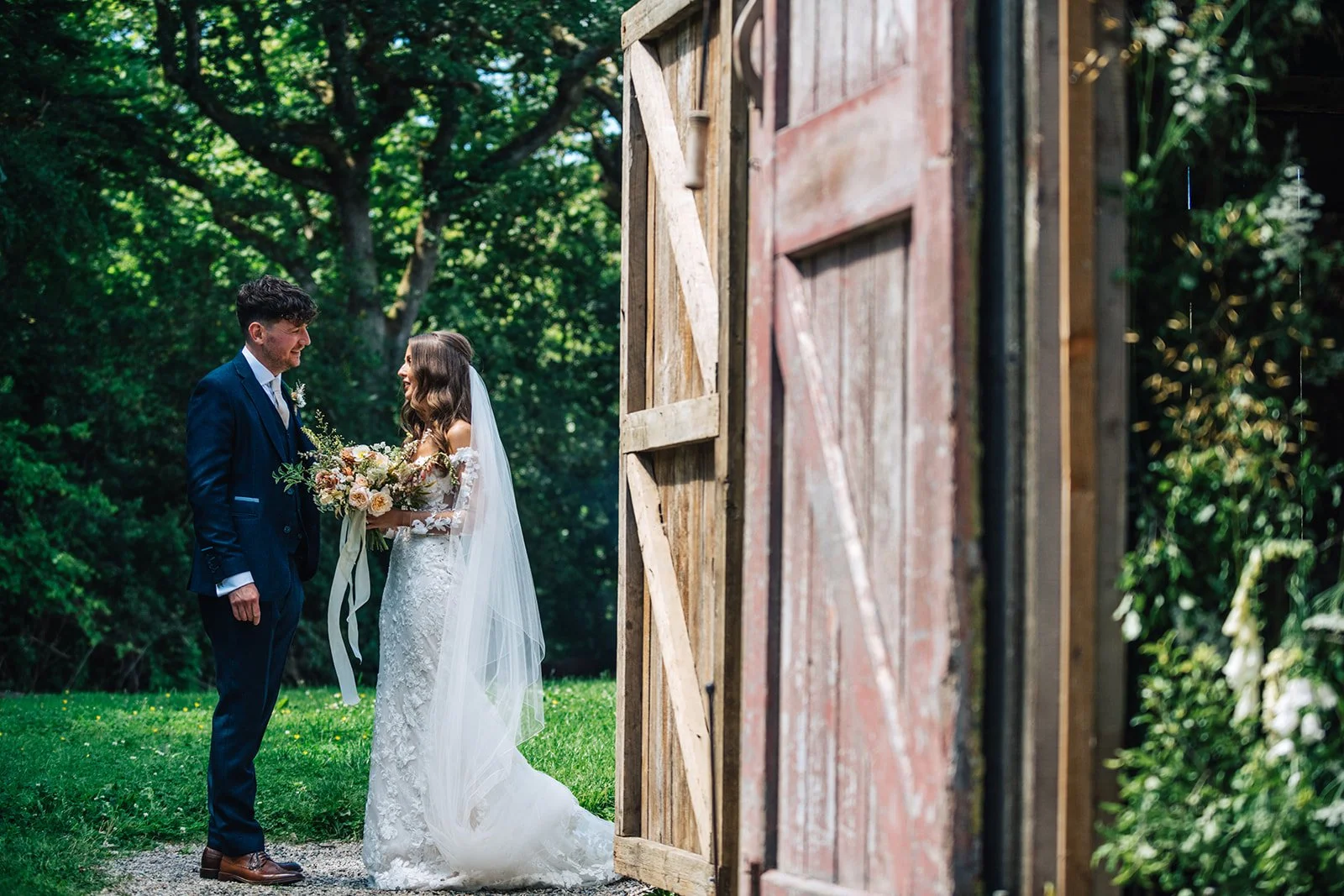 A bride and groom standing outdoors near a rustic barn door, sharing a moment on their wedding day surrounded by green trees.