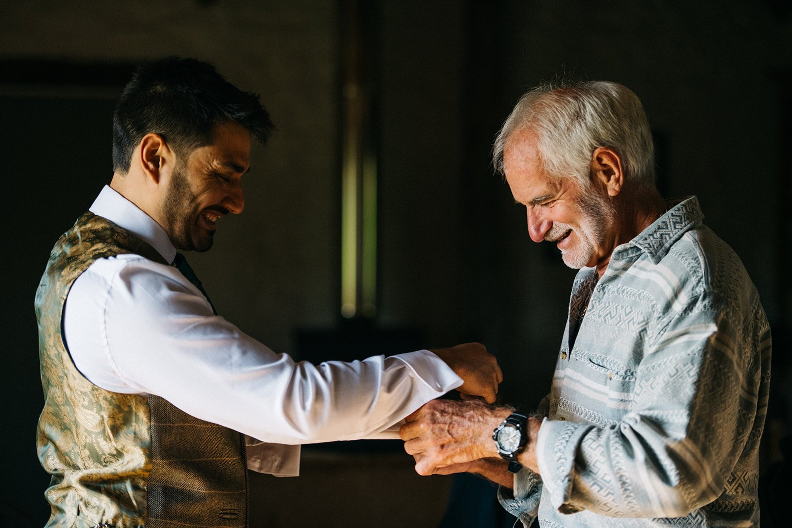 A young man and an older man share a joyful moment, smiling and holding hands, in a dimly lit room.