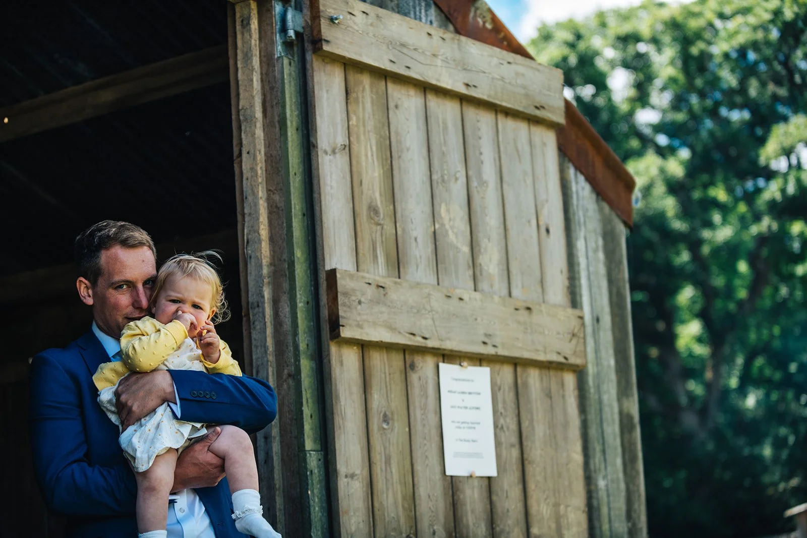A man in a blue suit holding a young girl with blonde hair, wearing a yellow sweater, outside near a wooden structure with greenery in the background.