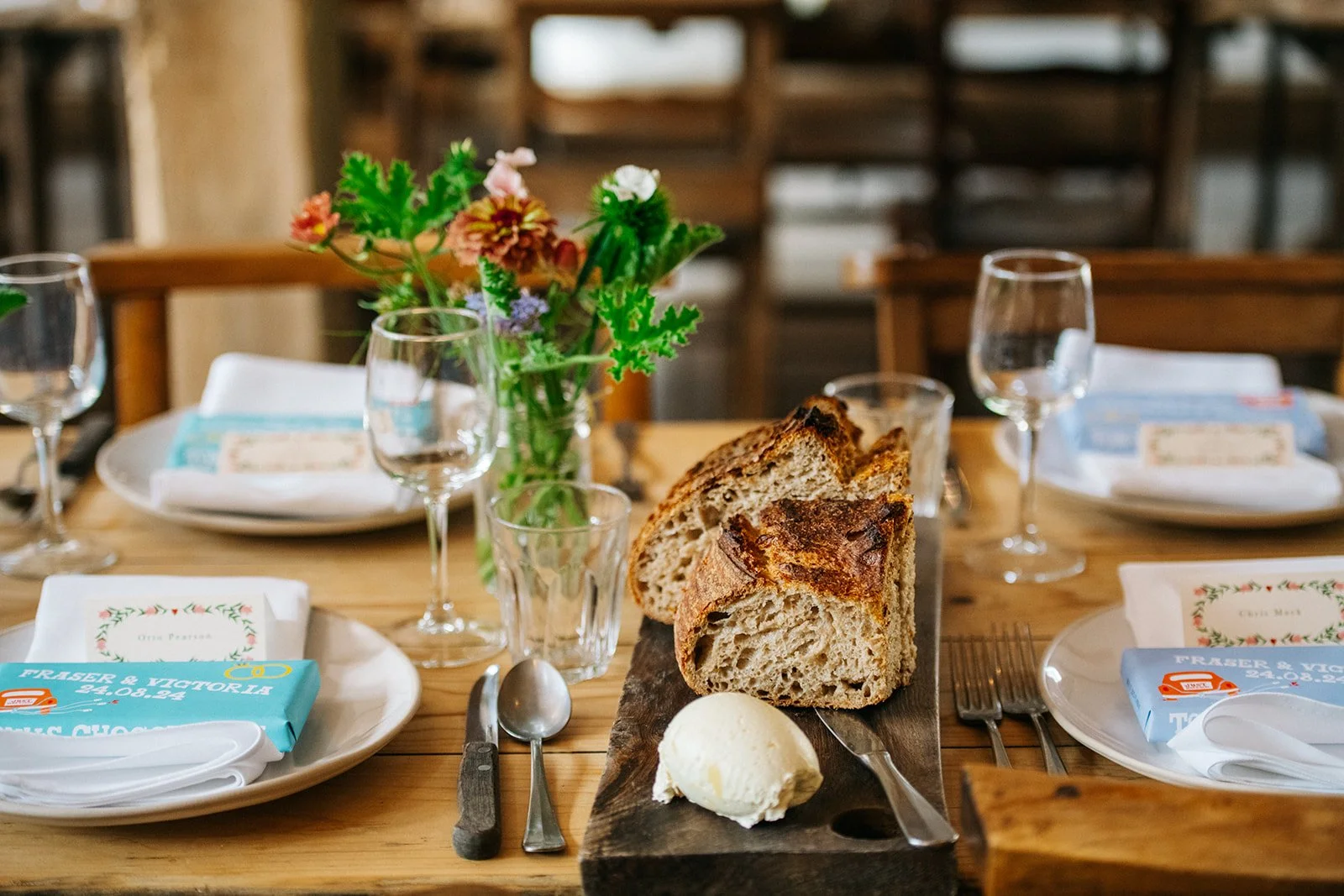 A rustic wooden table set for a meal with sliced bread, butter, and empty wine glasses, decorated with a small bouquet of colorful flowers in a glass jar.