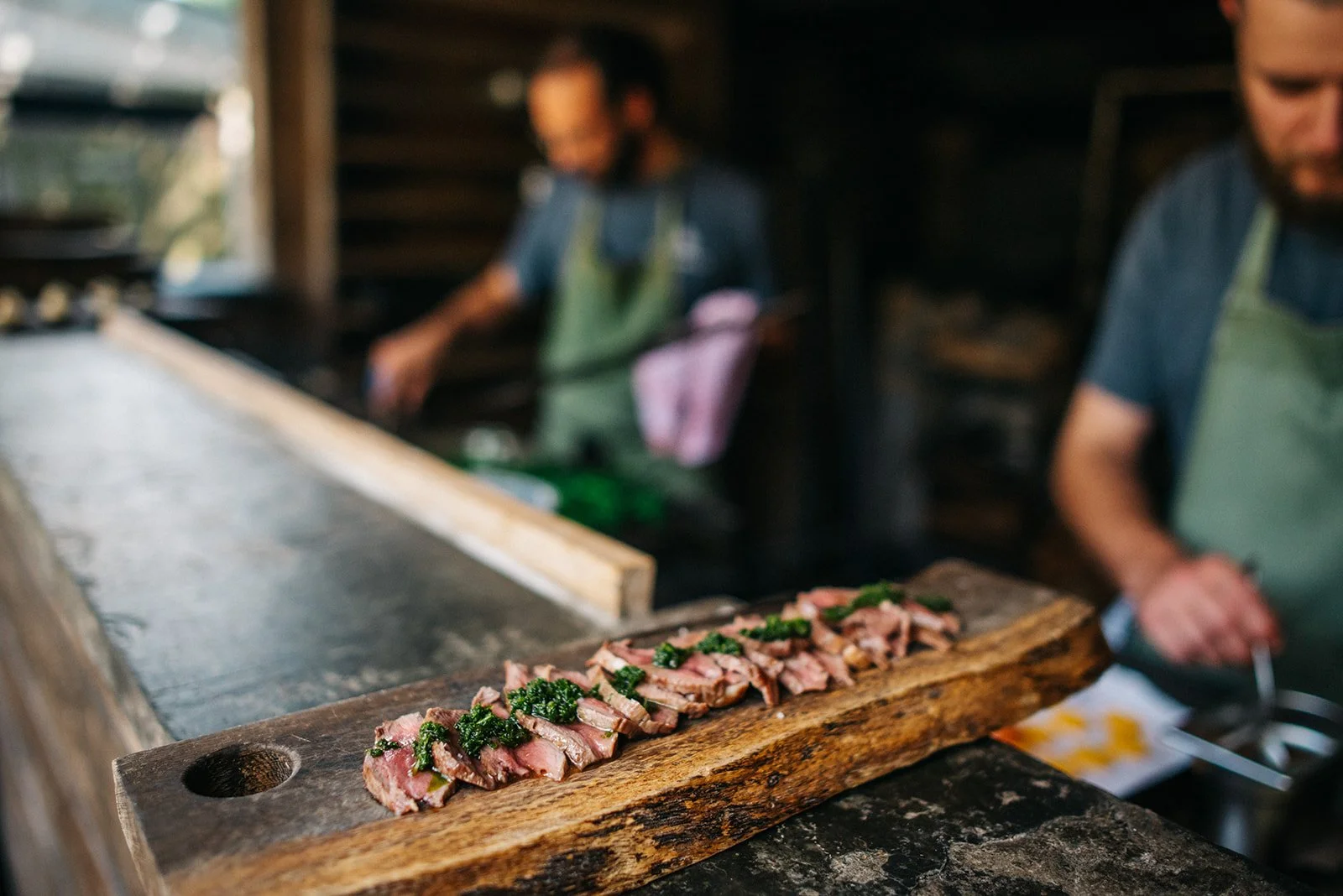 Sliced steak with green herb sauce on a wooden serving board in a rustic kitchen with two chefs in the background.