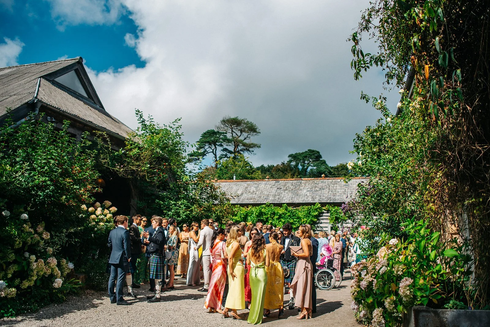 A group of people dressed in formal attire gathered outdoors in a garden on a cloudy day, with greenery and buildings in the background.