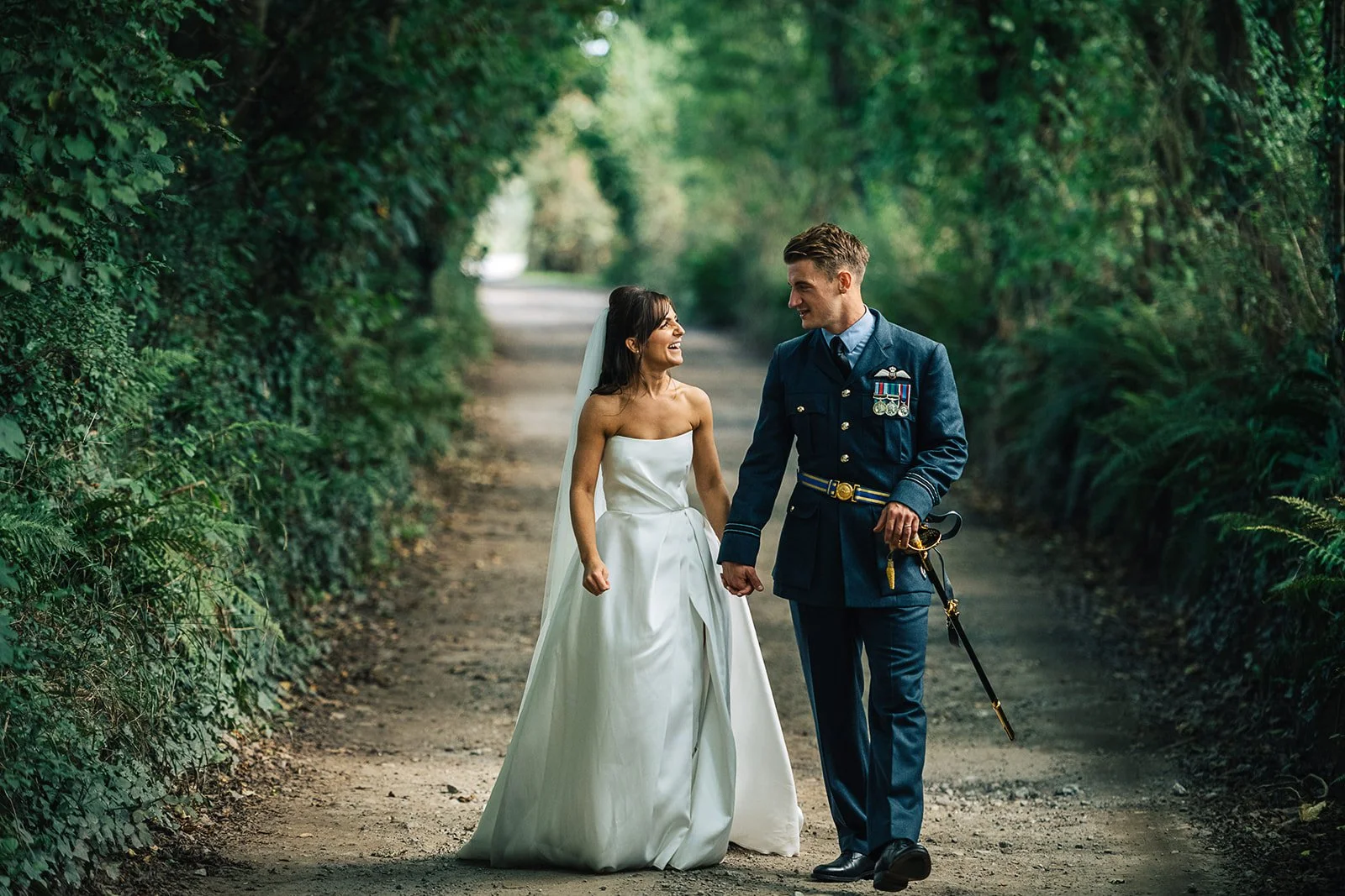 A bride and a groom, dressed in a wedding gown and a military uniform, walking together on a dirt path lined with green foliage, smiling at each other.