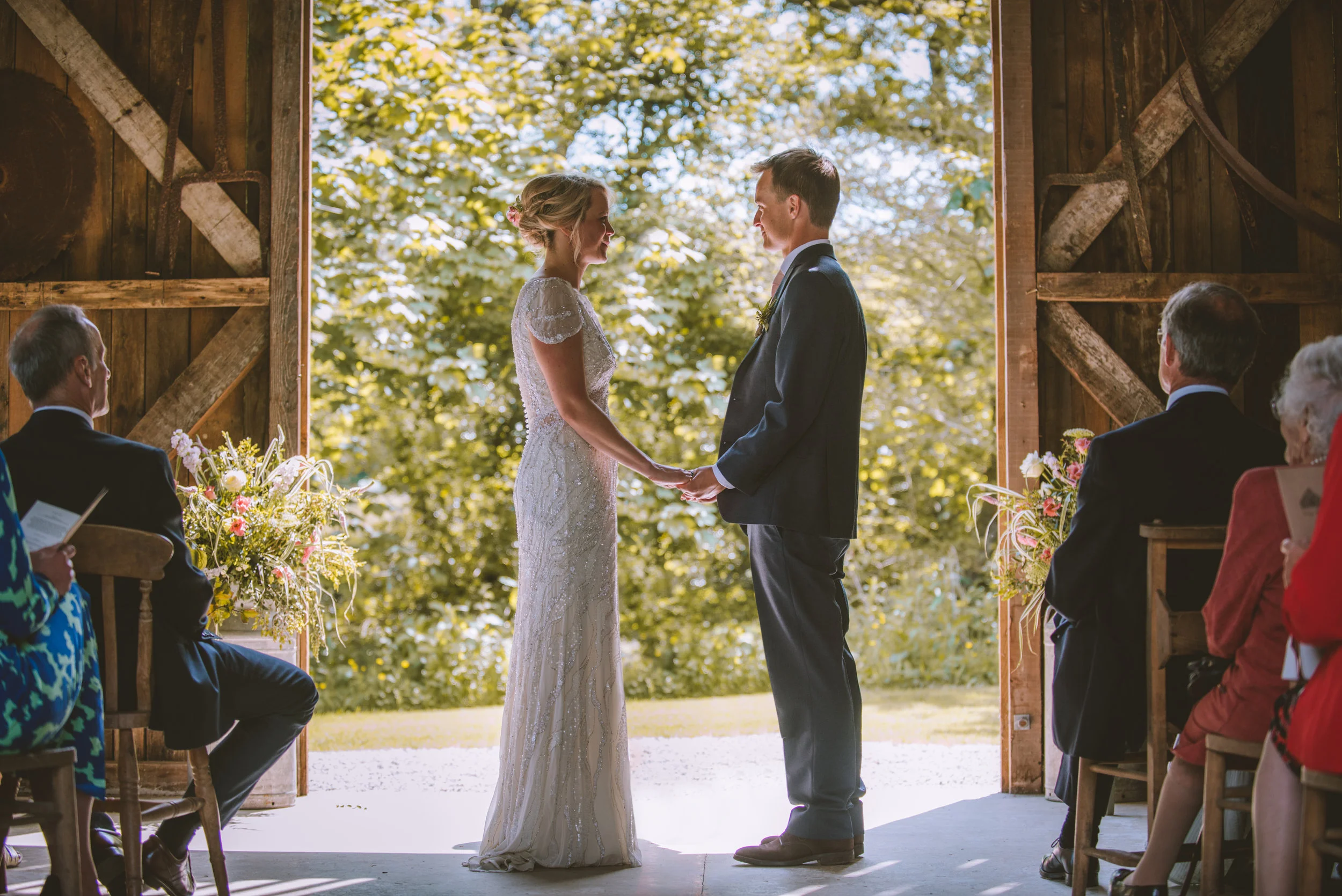 A bride and groom holding hands and smiling at each other during their wedding ceremony in front of a wooden barn door with trees behind them, surrounded by seated guests.