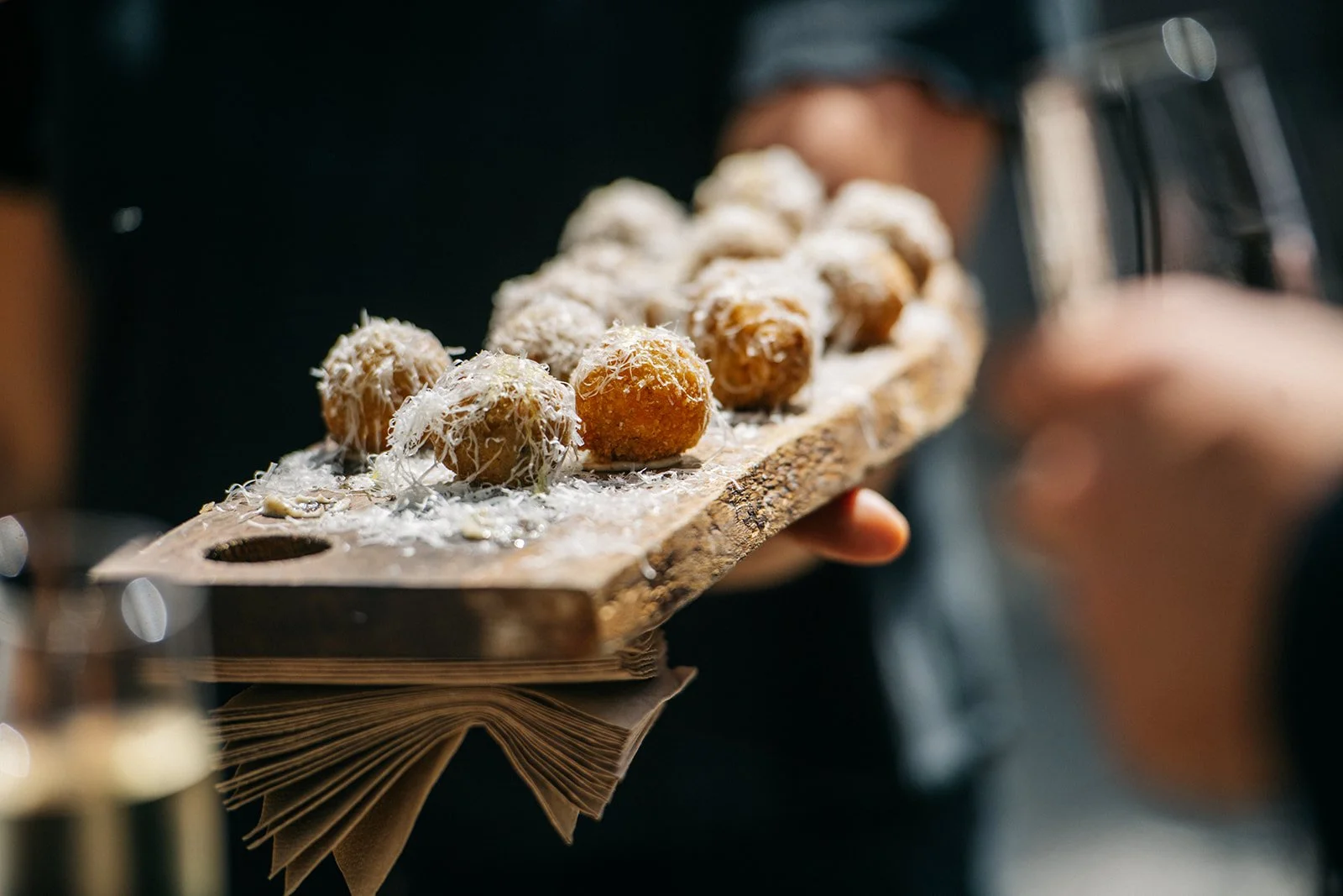 A person holding a wooden serving board with several dessert balls covered in shredded coconut, some on a piece of parchment paper.