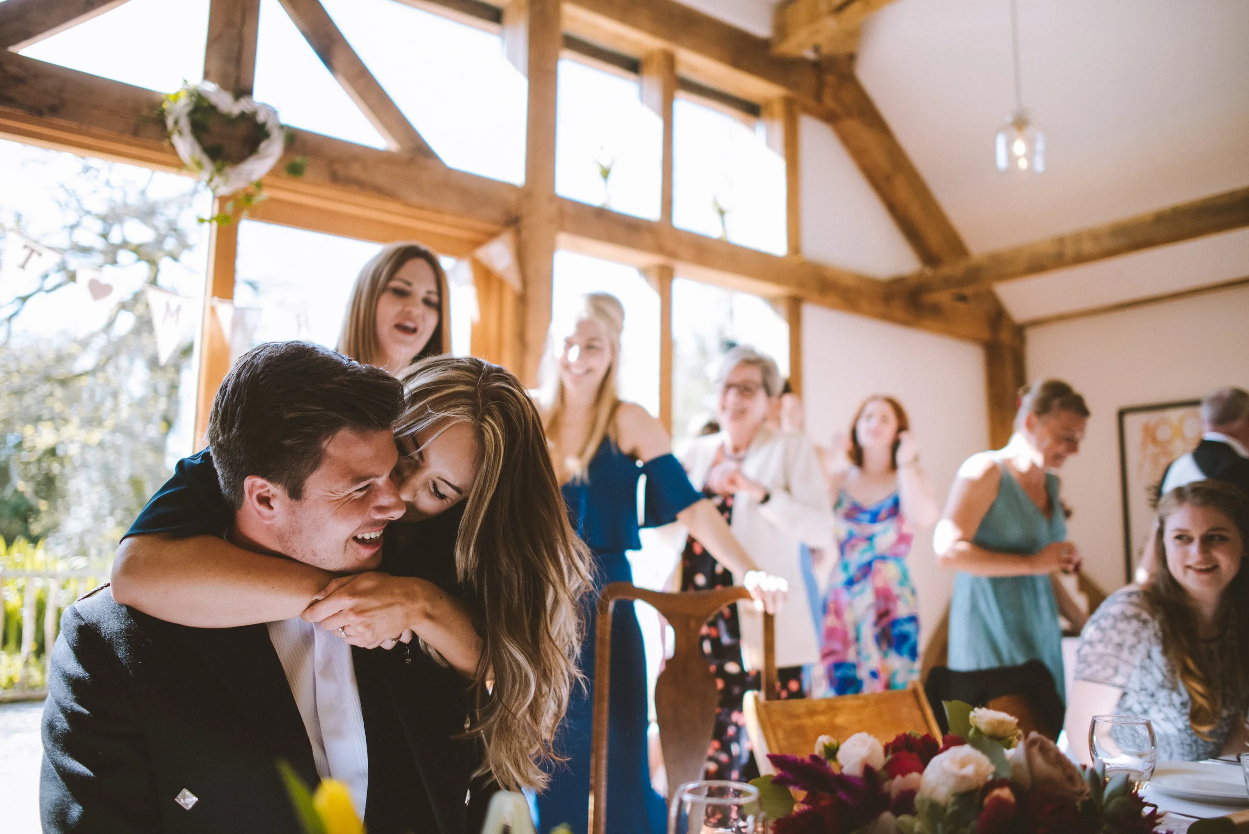 A joyful wedding reception with a groom and bride emulating a hug, surrounded by friends and family in a bright, wooden-beamed room with large windows, floral decorations, and a table with flowers and glassware.